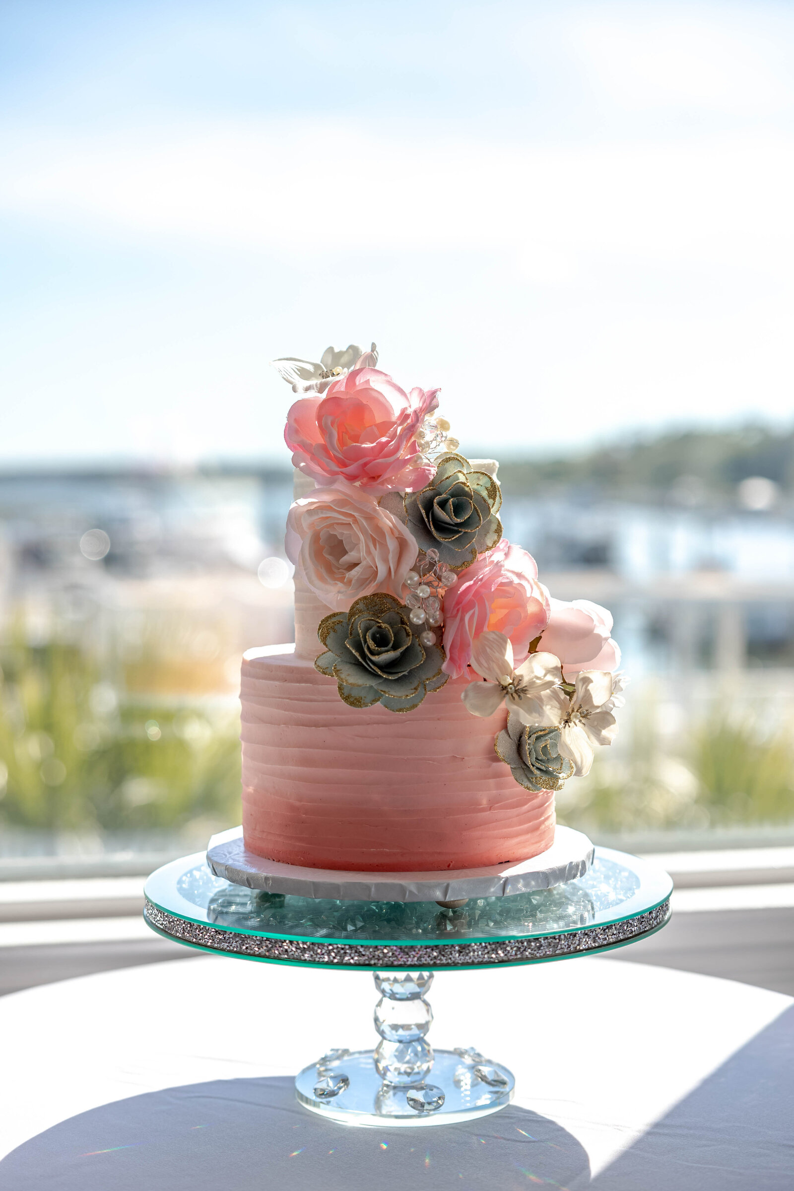 A stunning pink wedding cake with intricate floral details displayed on a glass stand by the water, captured at a Wrightsville Beach, NC wedding, blending elegance and coastal charm.