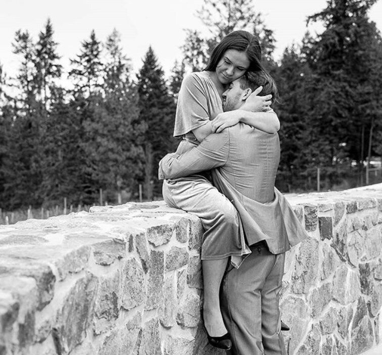 Couple sitting on a stone wall in a forest, sharing a cozy hug and smiling.