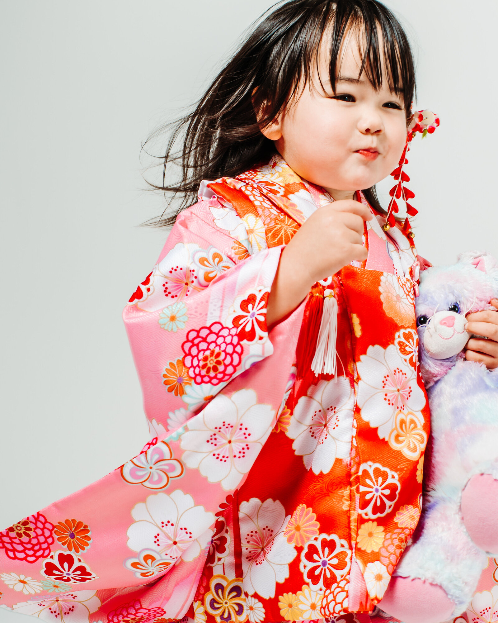Young girl in pink and red floral kimono holding pastel stuffed animal during Shichi-Go-San studio portrait in Los Angeles | 七五三 ロサンゼルス ピンクと赤の花柄着物の女の子 ぬいぐるみとスタジオ撮影