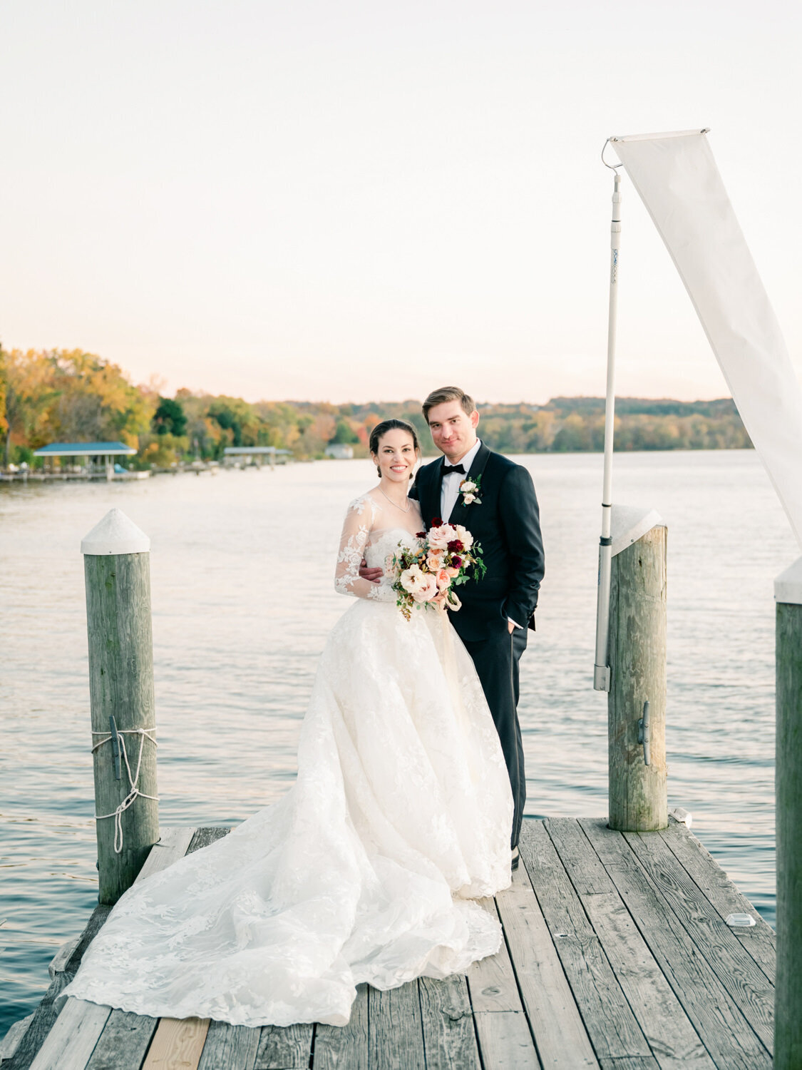 A fall wedding couple pose for the camera on a dock with the Finger Lakes behind them.