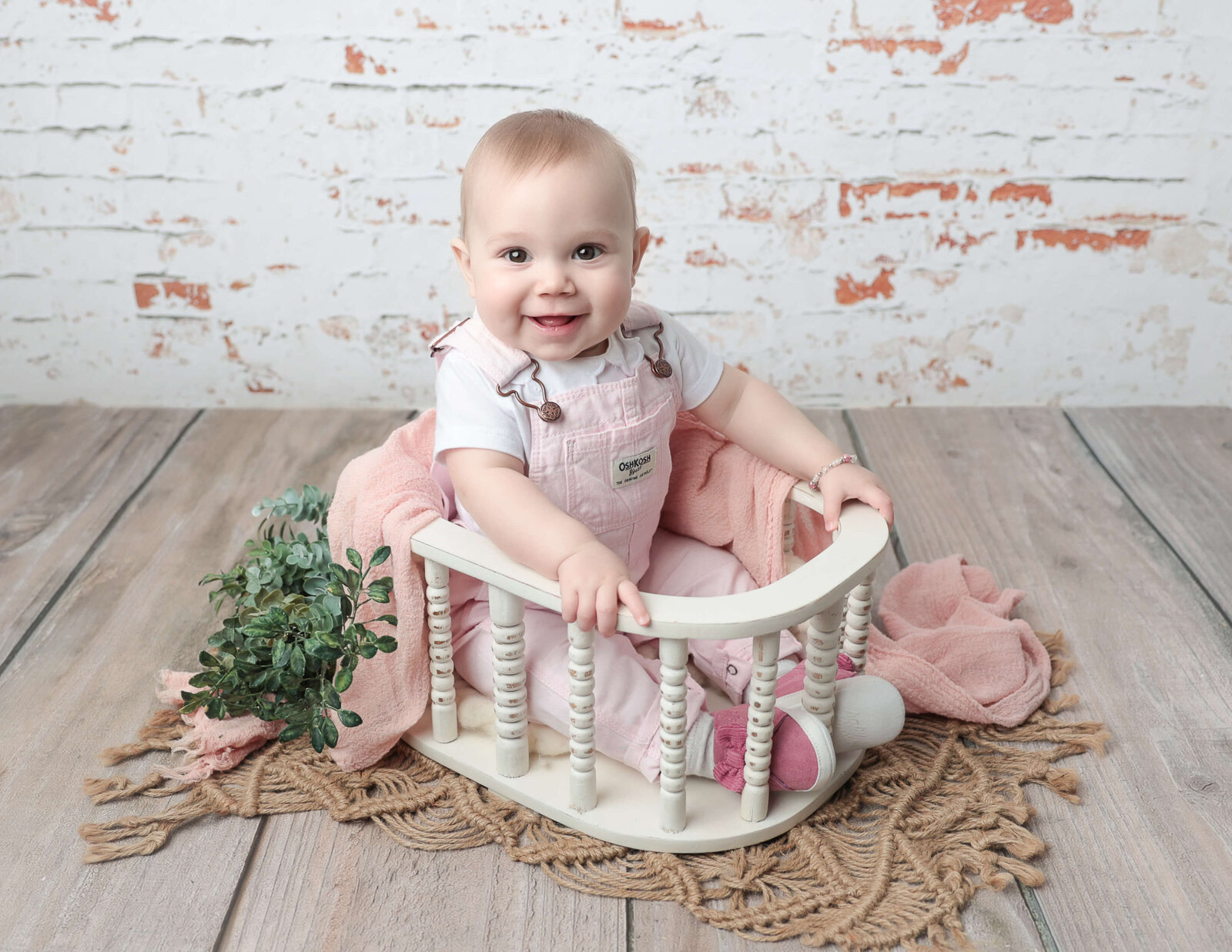 A joyful six-month-old baby sitting in a white basket during a milestone photography session in a studio in Rochester, NY, surrounded by soft blankets and greenery.