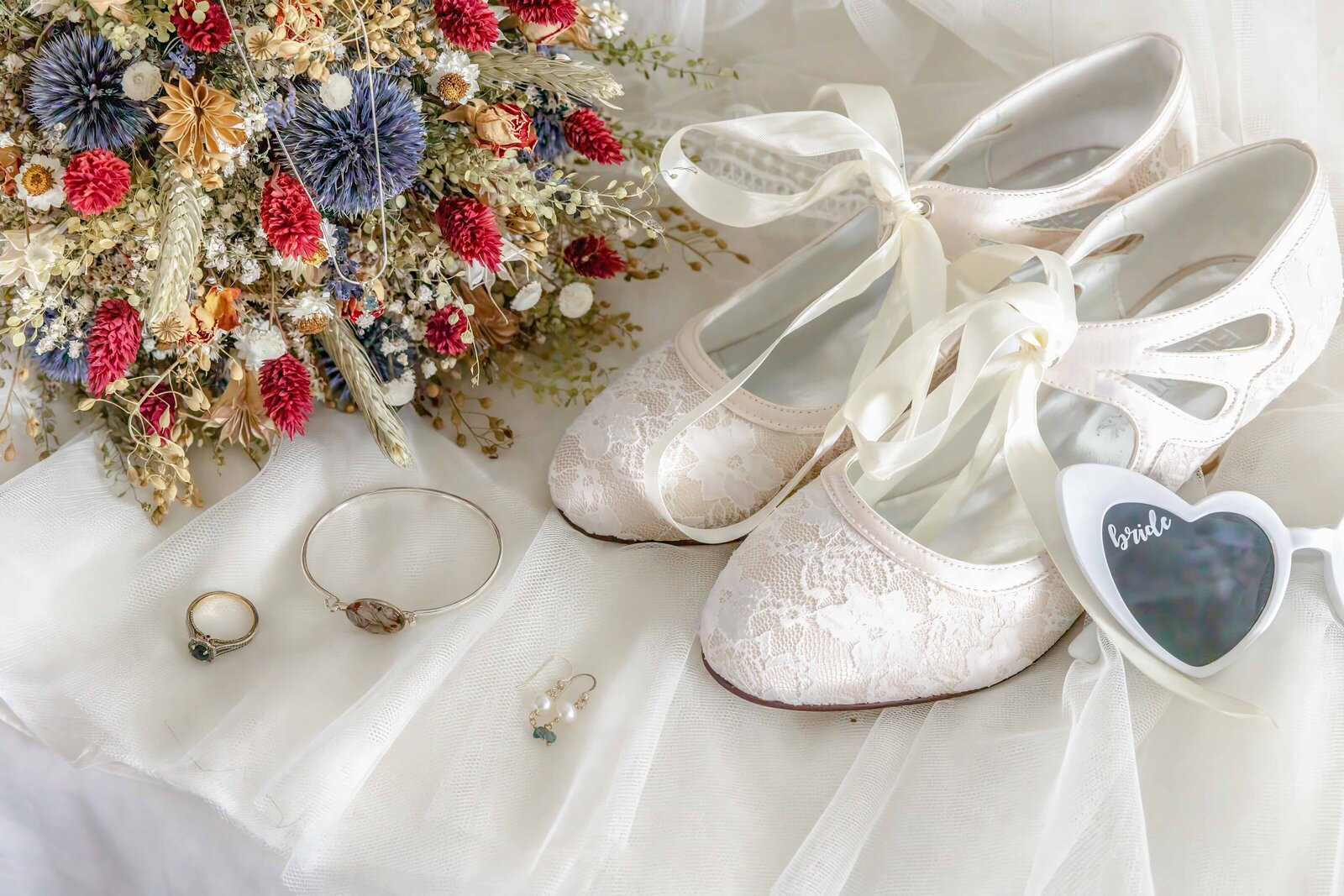 A bridal flat lay displaying lace wedding shoes, a dried floral bouquet, and bride sunglasses, captured at a Wilmington, NC wedding, blending elegant and rustic wedding details.