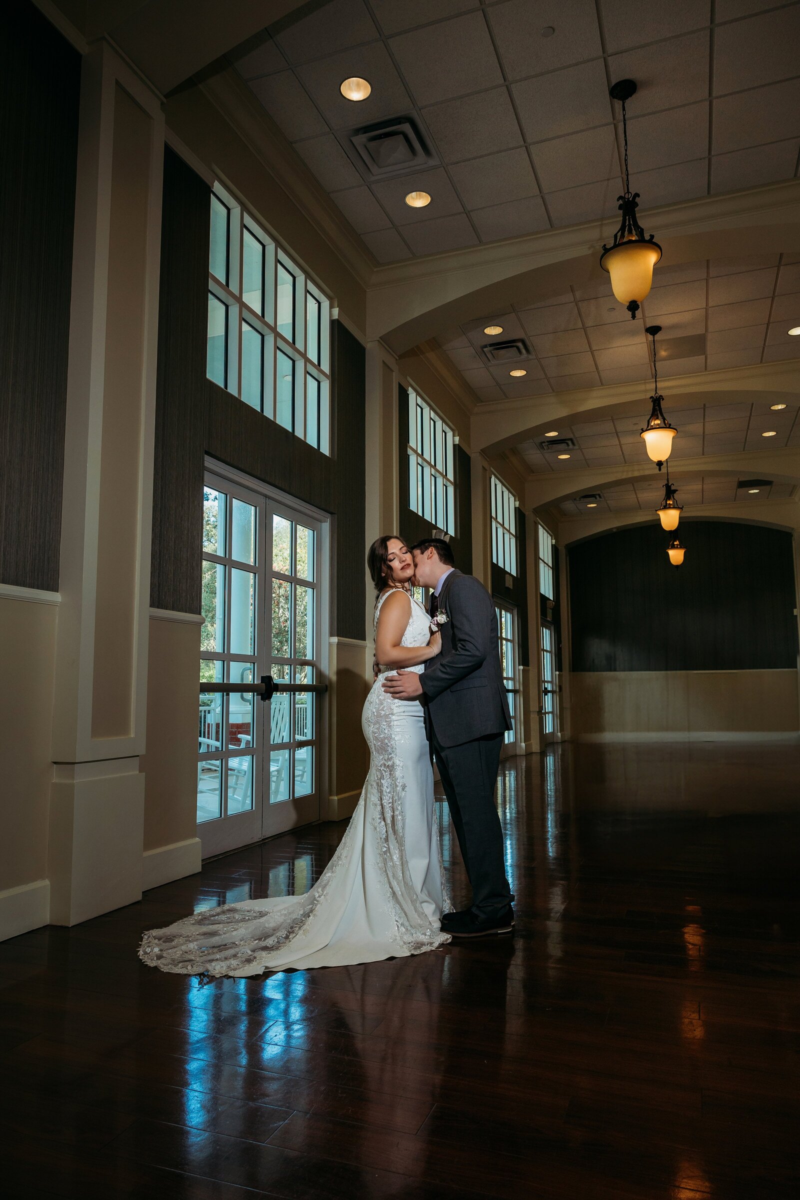 Bride and groom kiss in a grand hallway at Lake Mary Event Center, her lace gown cascading across the polished wood floor.