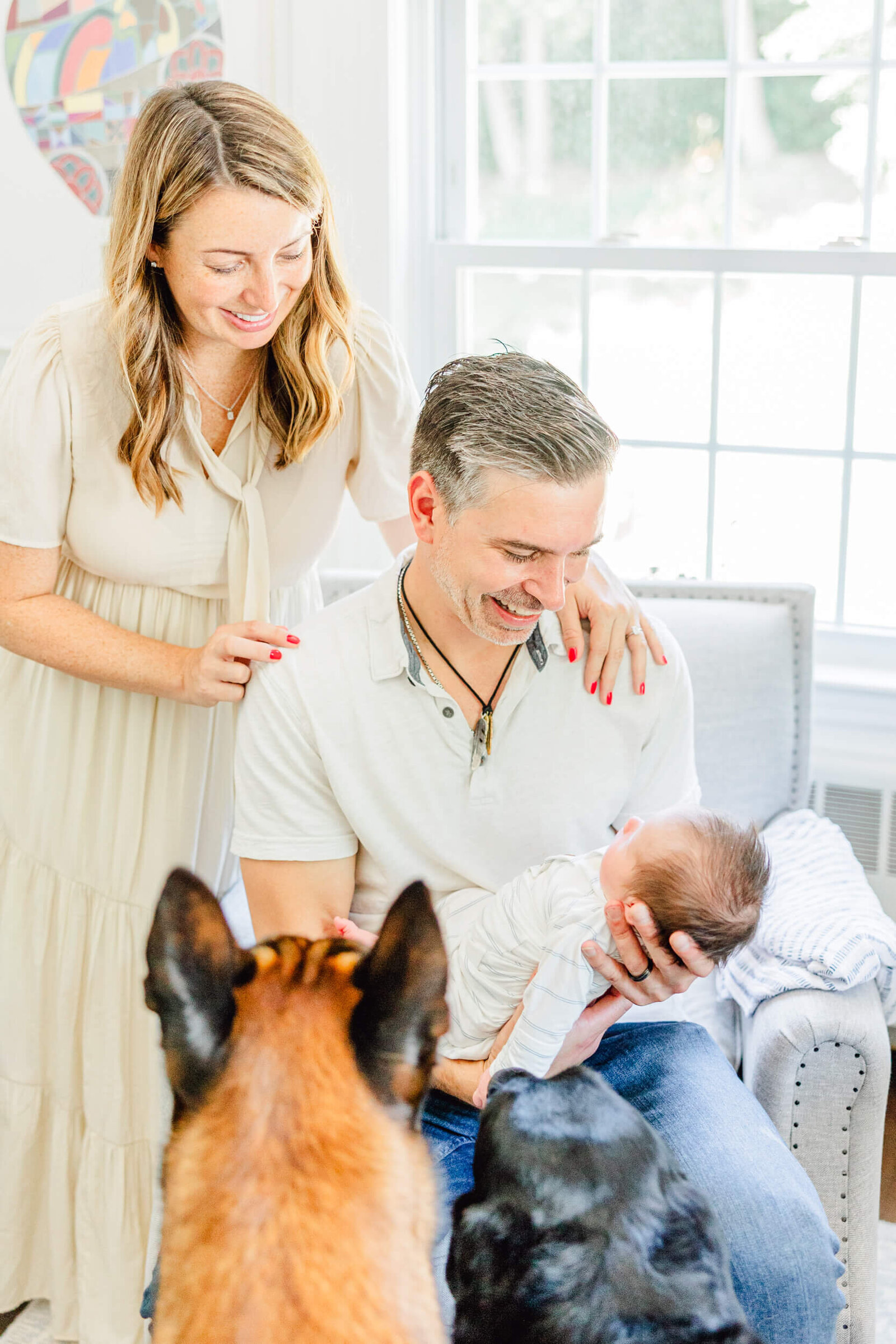 mom and dad smile at baby while the back of the heads of two dogs poke into the frame