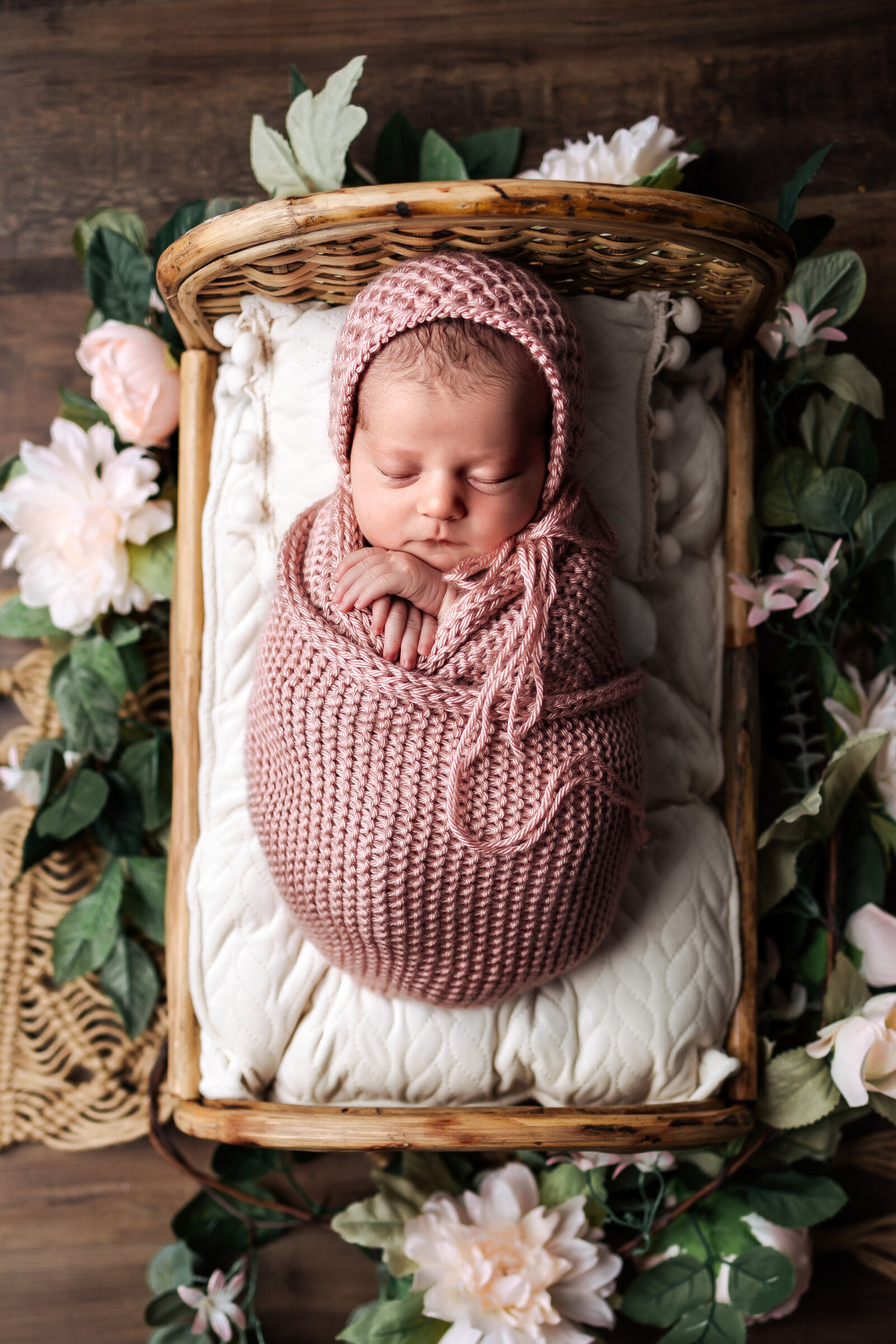 Newborn girl wrapped in pink with bonnet laying in tiny bed with flowers