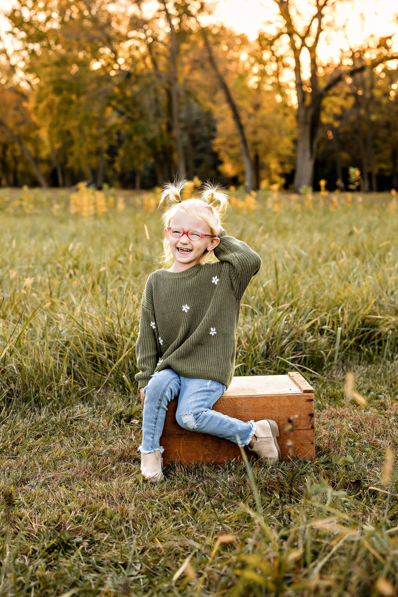 A young child with blonde hair and pigtails sits on a wooden box in a grassy field, capturing the essence of outdoor portraits. They're wearing a green sweater with white flowers, blue jeans, and beige shoes. The child smiles amidst trees with autumn leaves in the background.