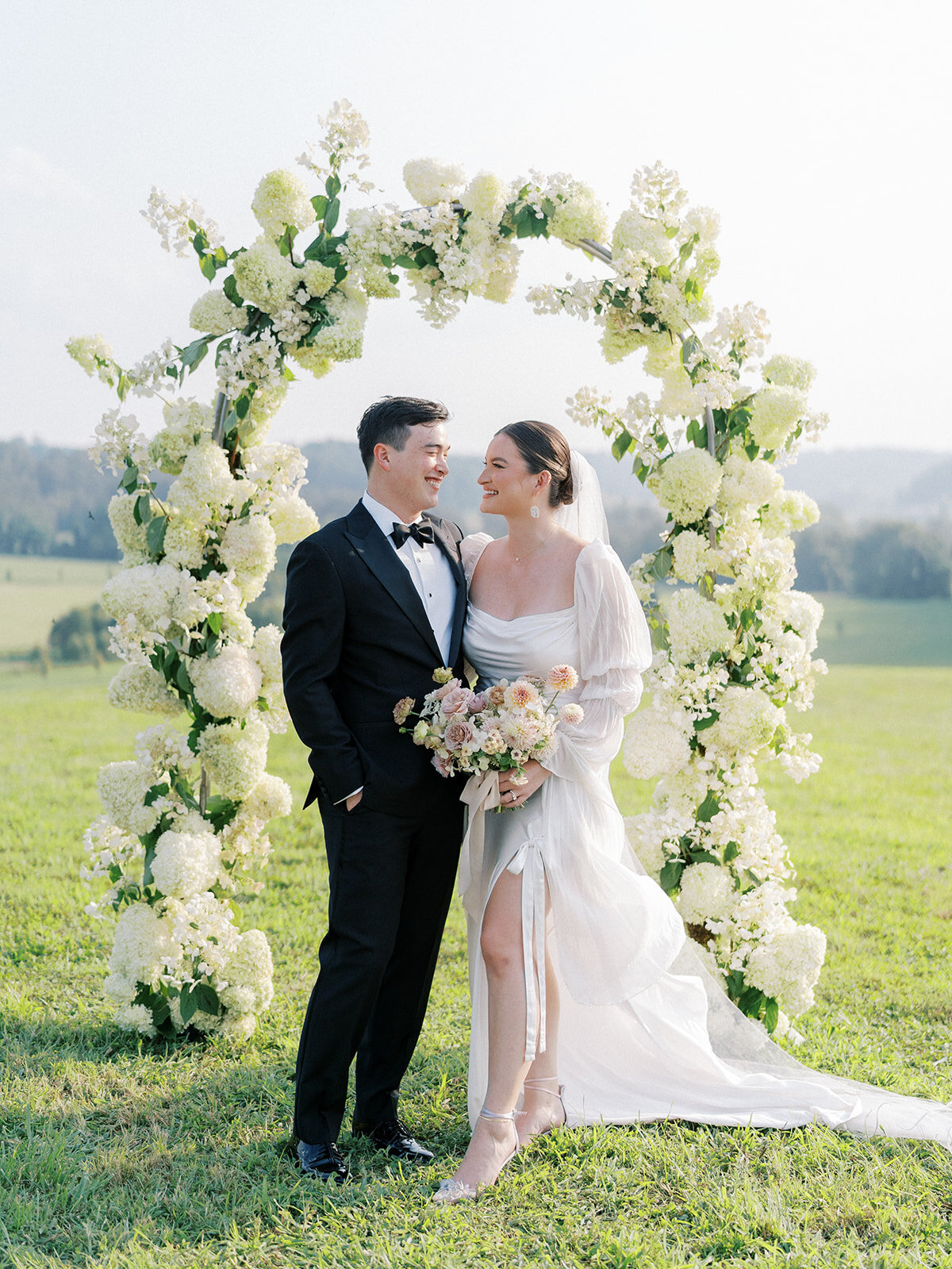Portrait of bride and groom under a full coverage floral arch filled with white airy quick-fire hydrangea and limelight hydrangeas.