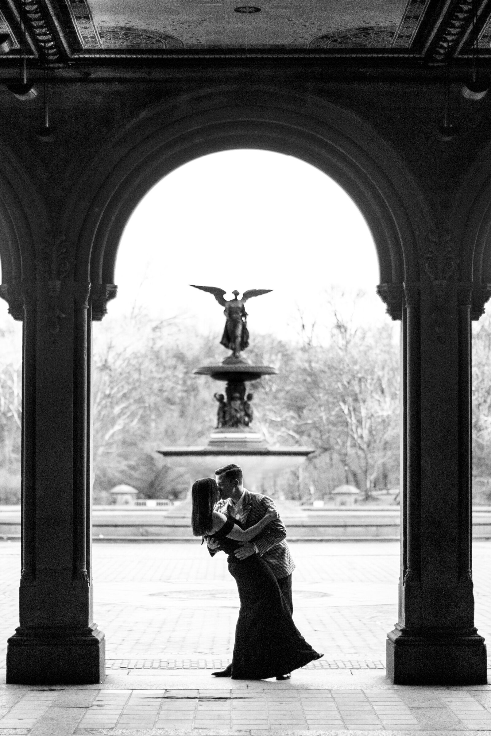 A couple embraces under an arched structure, silhouetted against a fountain with an angel statue in the background. The scene is black and white, creating a romantic and timeless atmosphere.