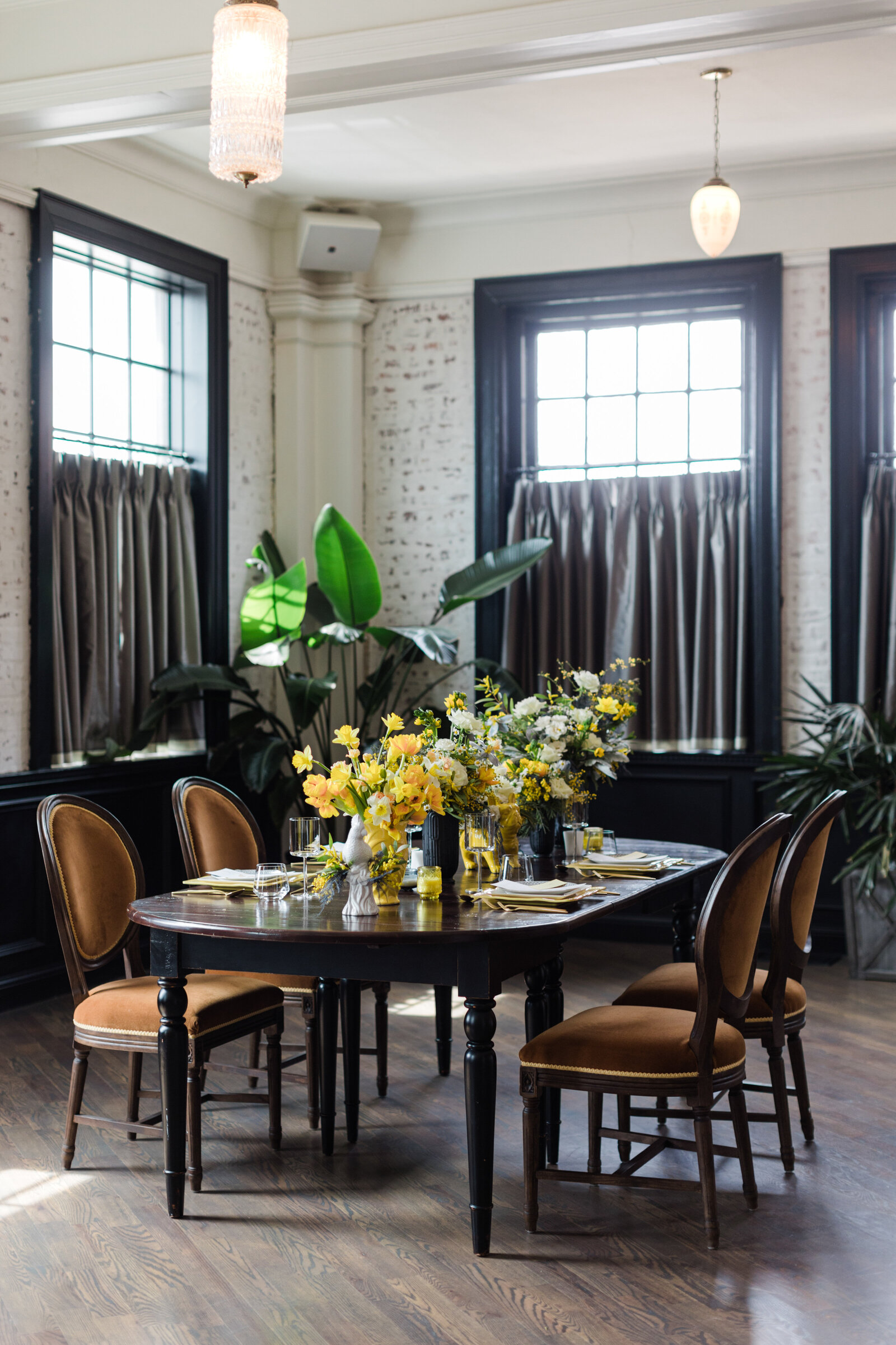Detail shot of a reception table at the Mason in Dallas, Texas. The dark wooden table is topped with multiple bouquets of white and yellow flowers and each seat has a place setting surrounded by cushioned dining chairs.