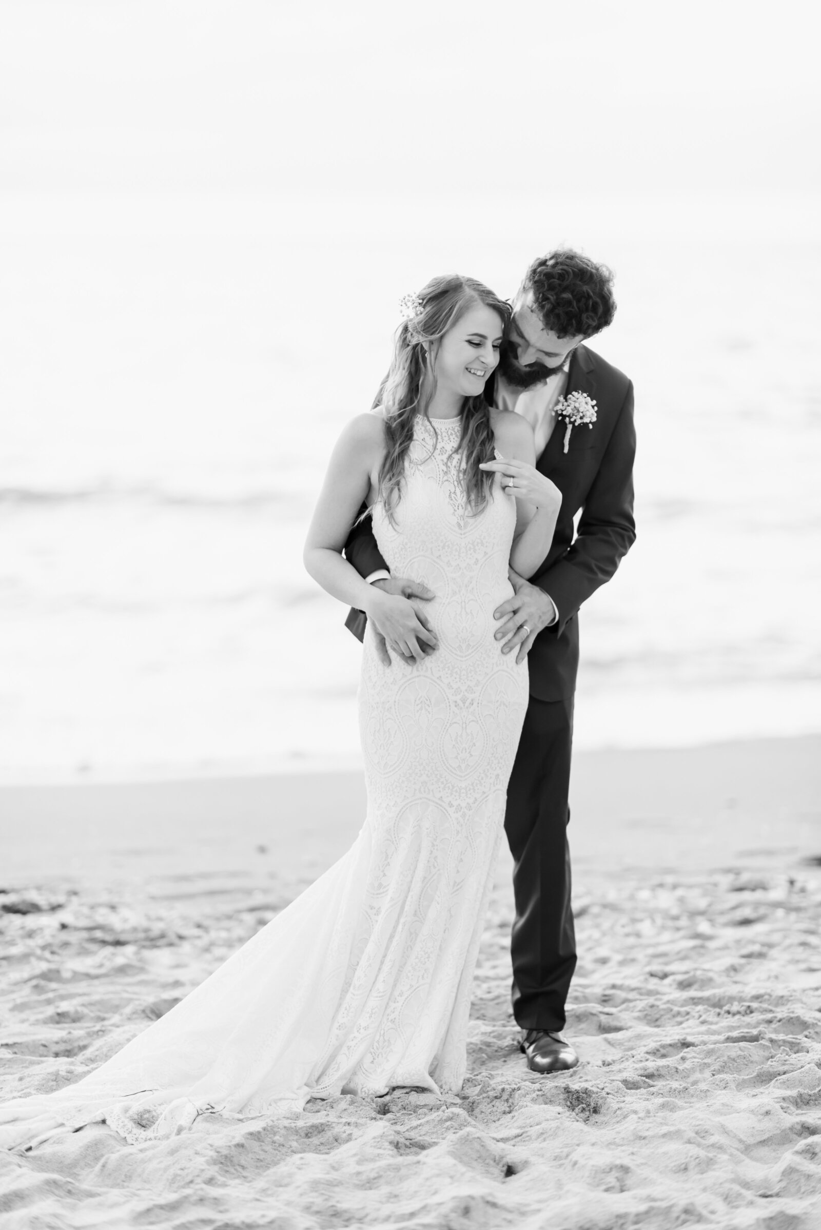 black and white photograph of bride and groom on the beach. She is standing in front of him and he is leaning in to kiss her cheek