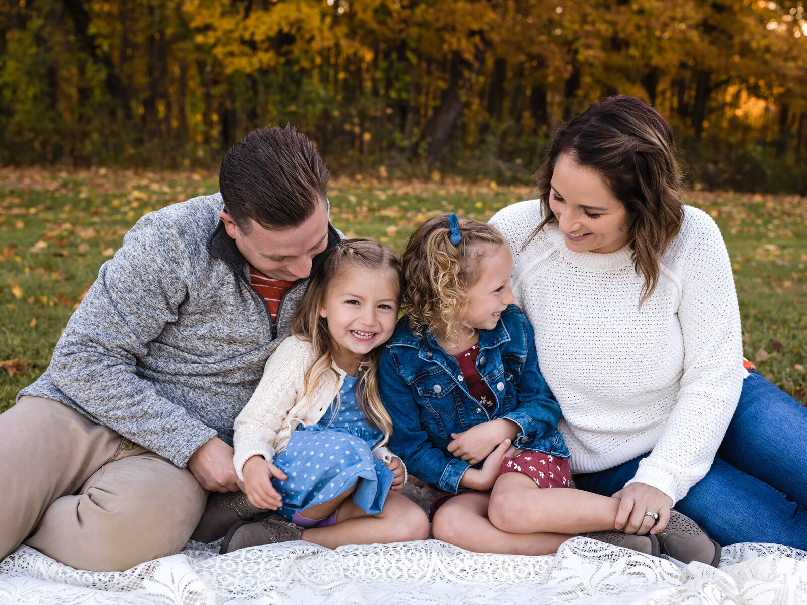 mom and dad sitting with two daughters on white lace blanket for fall family photos