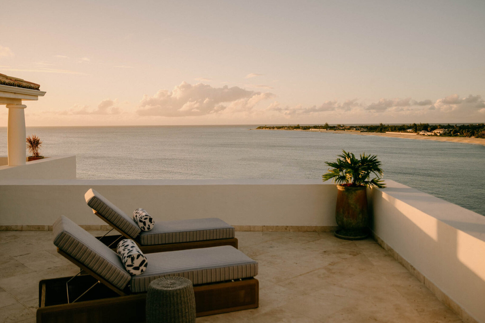 Sunset view from a coastal terrace with two cushioned lounge chairs and a small side table. A large potted plant is on the corner of the stone-tiled area, overlooking calm waters and distant coastline under a partly cloudy sky.