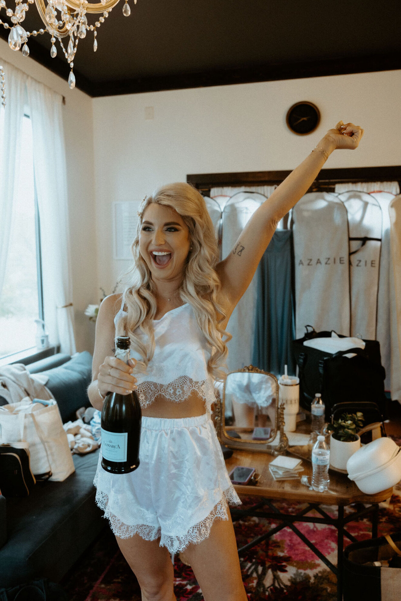 In a room adorned with dresses and bags, a woman in a white lacy outfit radiates joy, holding a champagne bottle. This bridal beauty experience captures the delight of what to expect from a wedding makeup artist in Texas.