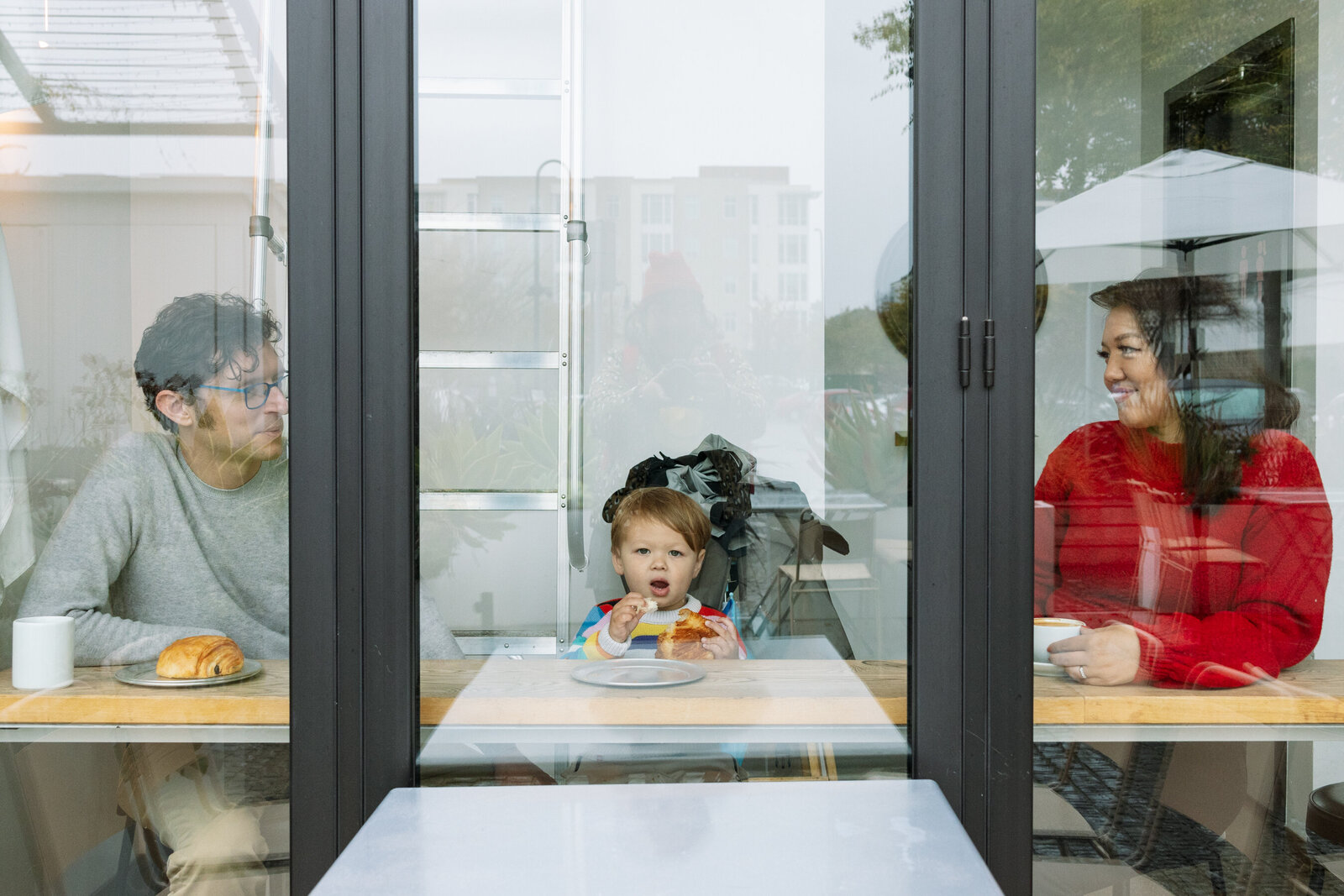 photo taken through window of a coffee shop of a family of three with toddler son in the middle