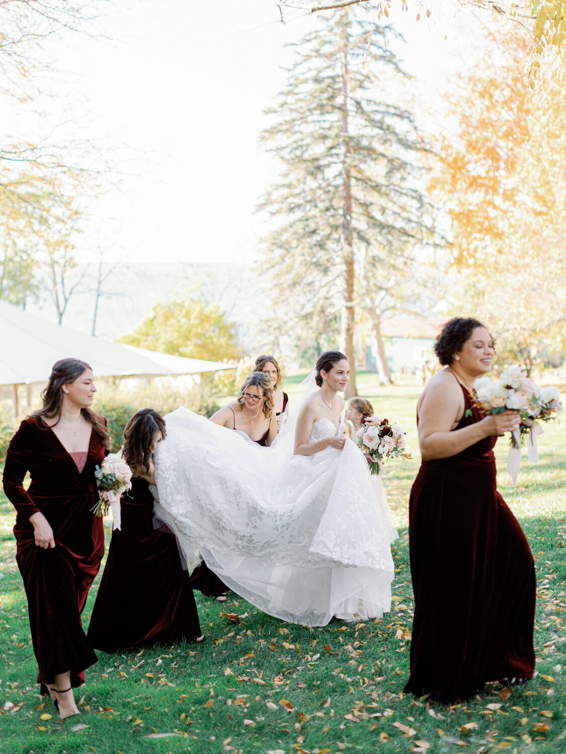 Bridesmaids in maroon dresses walking with bride in the fall leaves of New York resort.