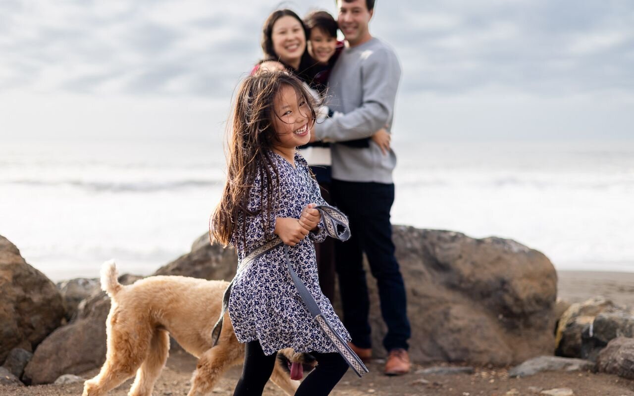 Young girl laughing and running with her dog while her family smiles behind her by the ocean – Ellobelle Photography – Bay Area Family Photographer