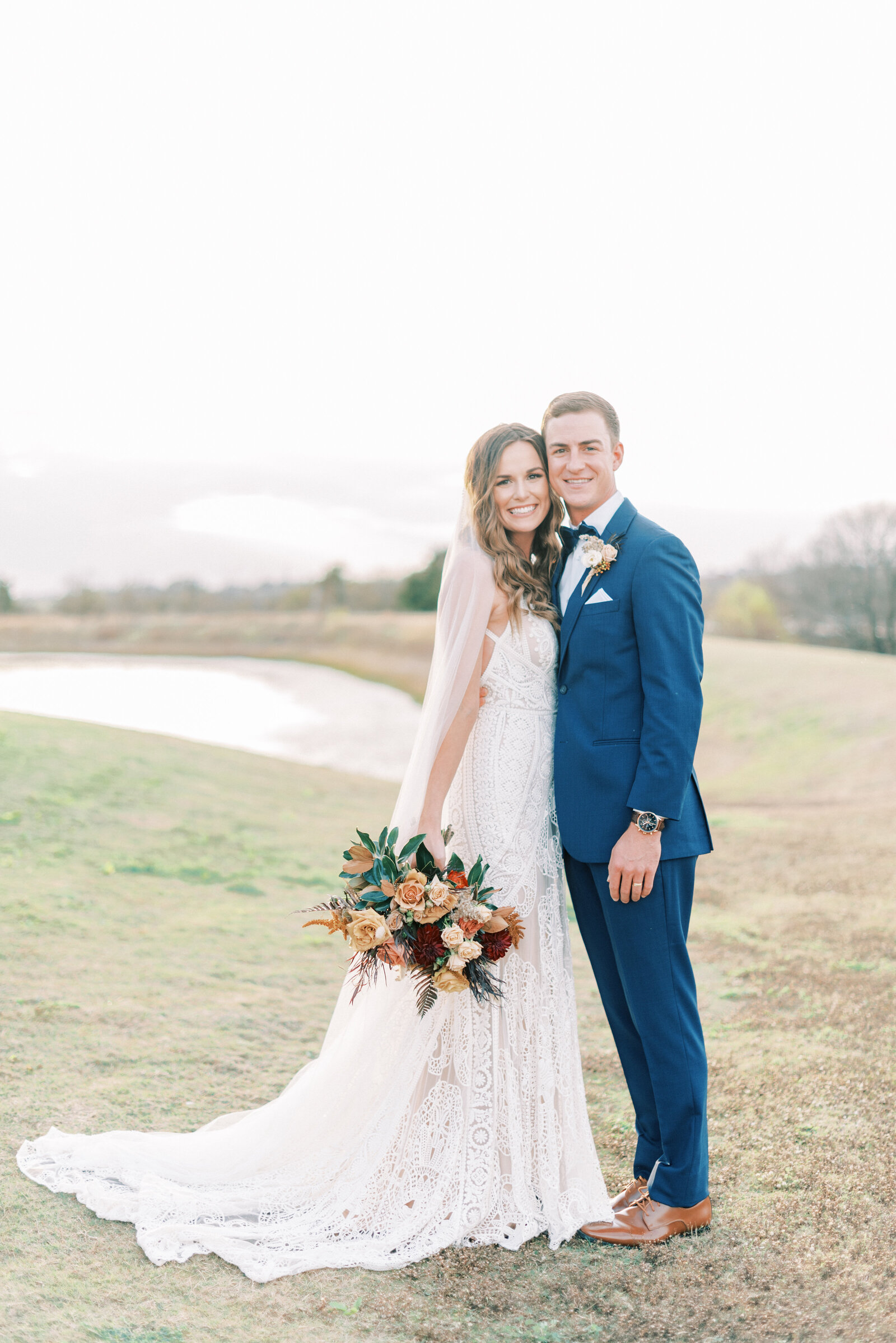 Portrait of a bride and groom in a white lace wedding gown and blue suit holding a bouquet on a field.