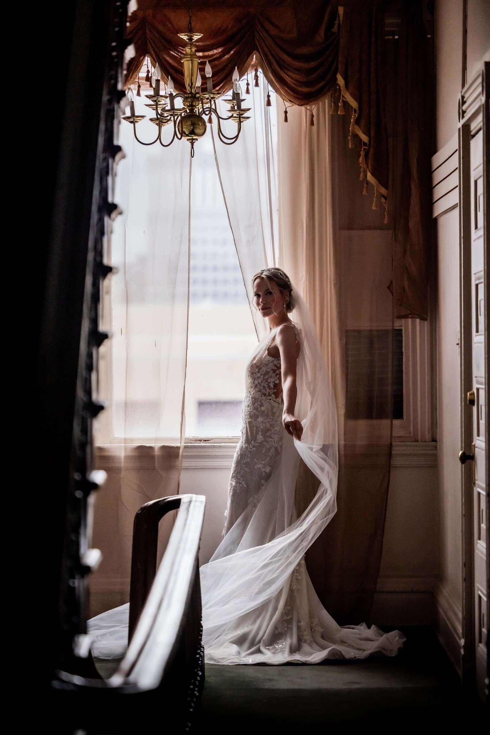 Capturing the essence of bridal elegance, this stunning couture portrait features a bride framed by a window's natural light. The delicate interplay of light and shadow highlights the intricate details of her gown, creating a serene and ethereal atmosphere. This image is perfect for those seeking inspiration for their own bridal look or anyone who appreciates the art of fine wedding photography.