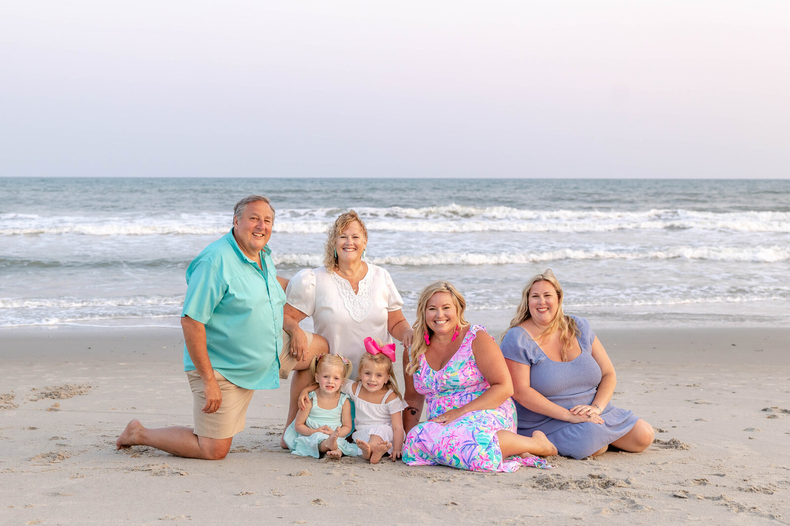 A multi-generational family sits on the sandy shore with ocean waves behind them. Dressed in colorful summer attire, they share joyful smiles, ideal for professional family photography in NC, capturing family warmth and connection.