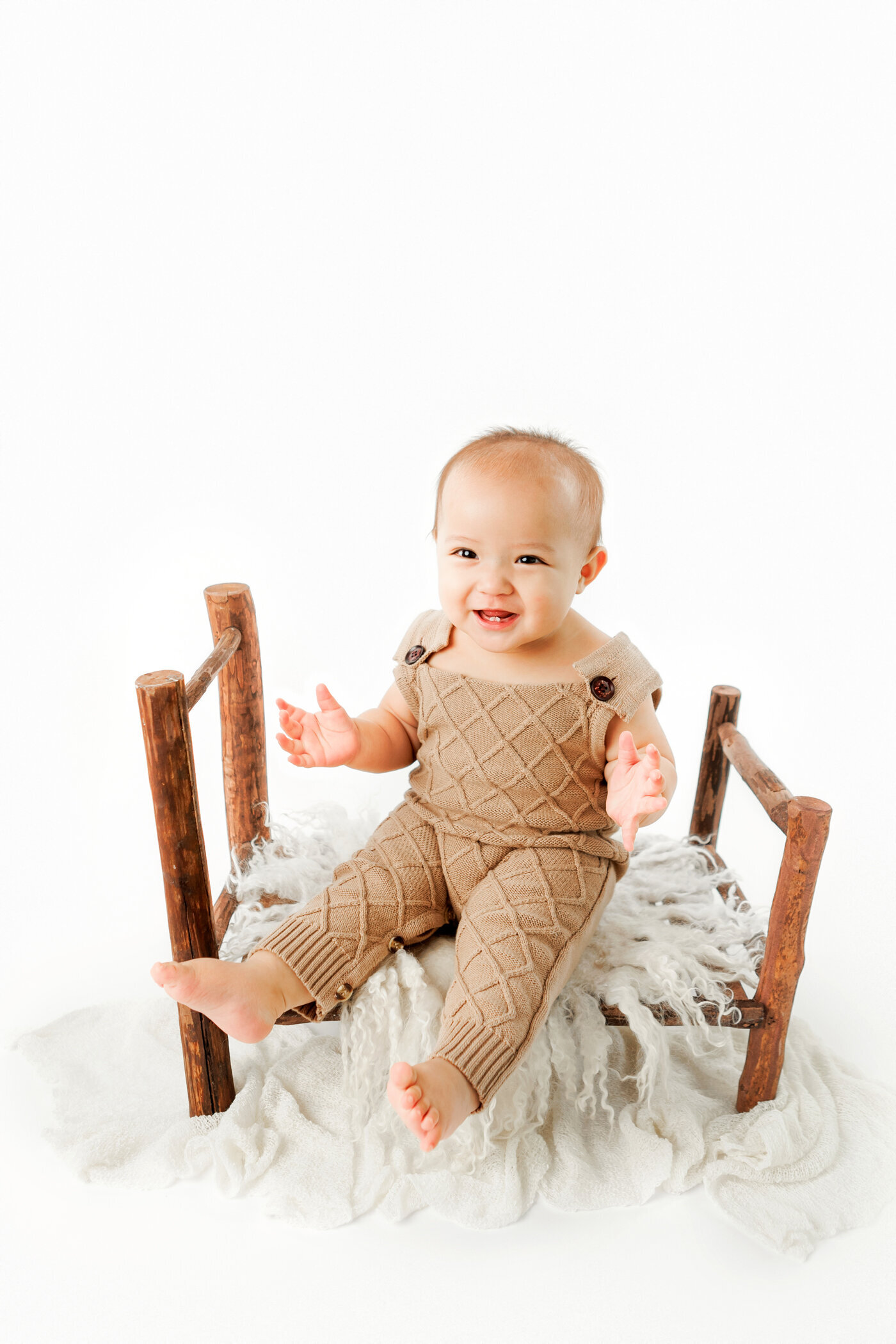 Baby boy sitting on wooden toy bed. Baby is wearing a brown romper sitting on neutral layers. 