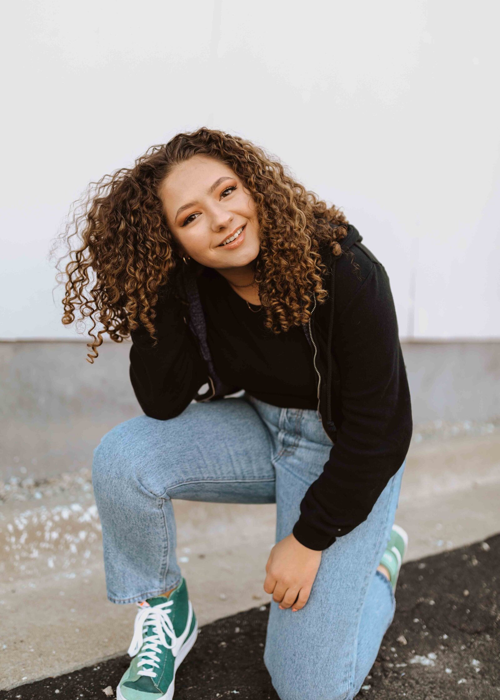 Girl kneeling down with hand in her curly brown hair. Smiling and looking at the  at the camera.
