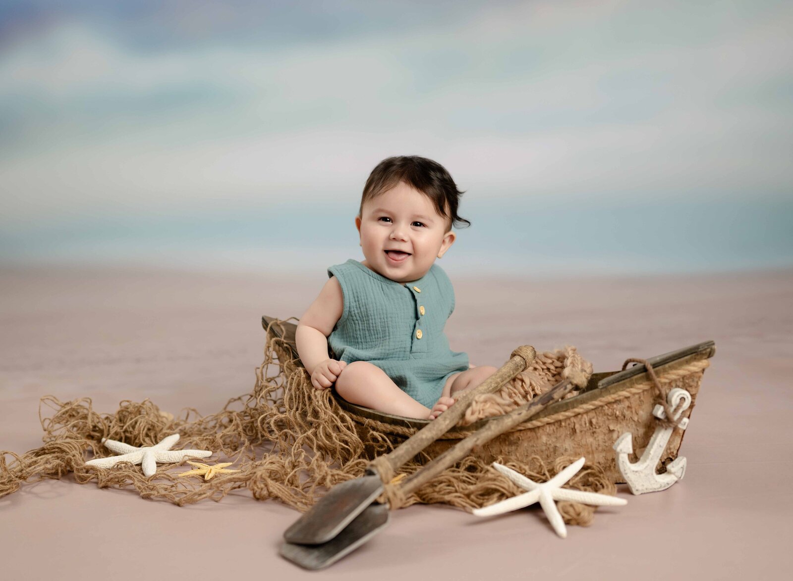 Baby boy sitting in a wooden boat by ocean backdrop with a big smile.