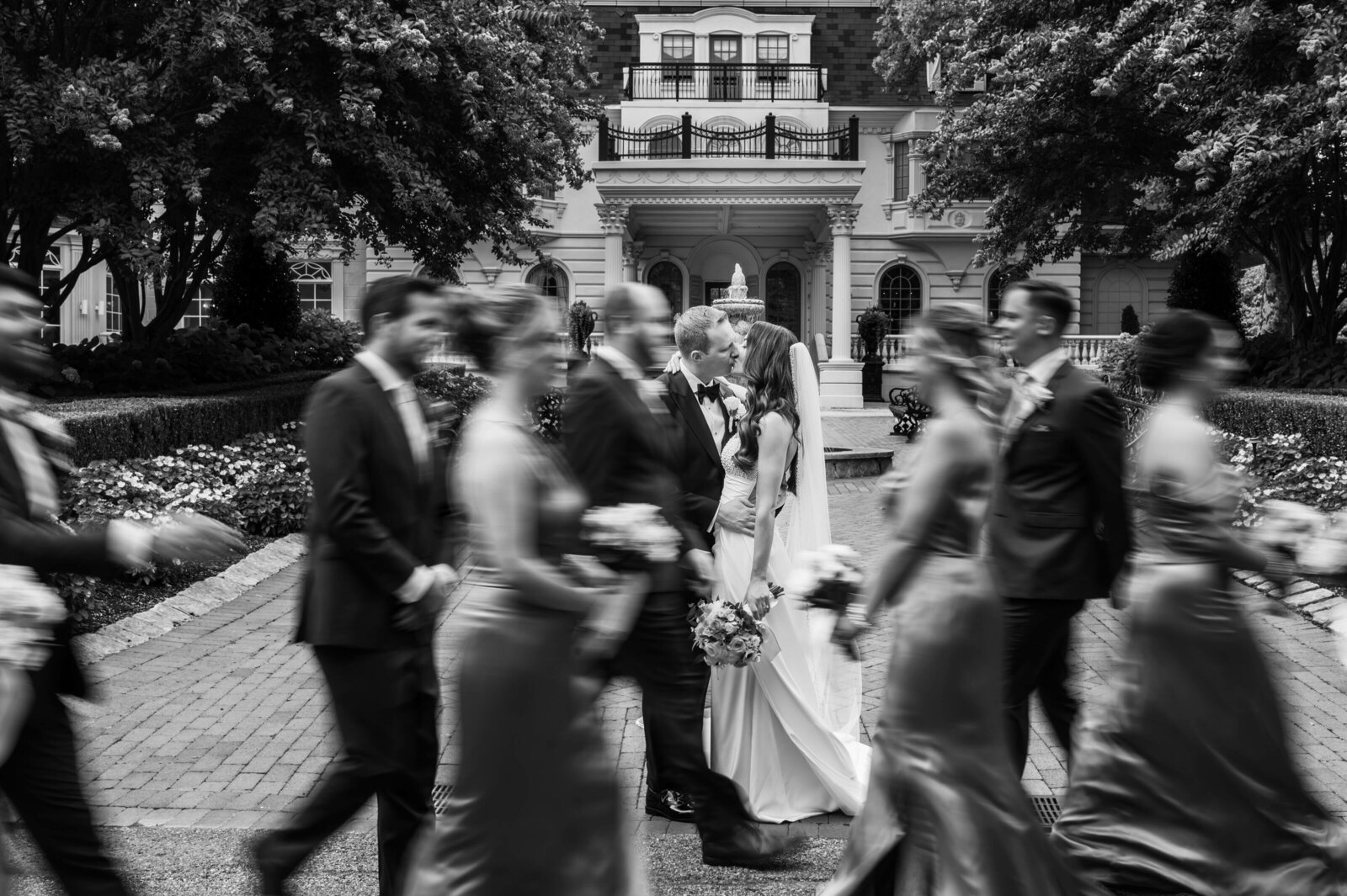 A bride and groom kiss in the center, surrounded by their wedding party walking past in a blur, with a large building and trees in the background.