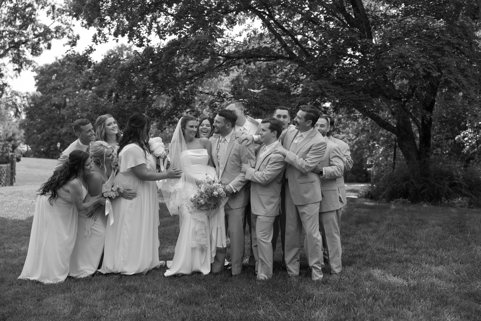 Wedding party hugging bride and groom with trees and bushes in the background.