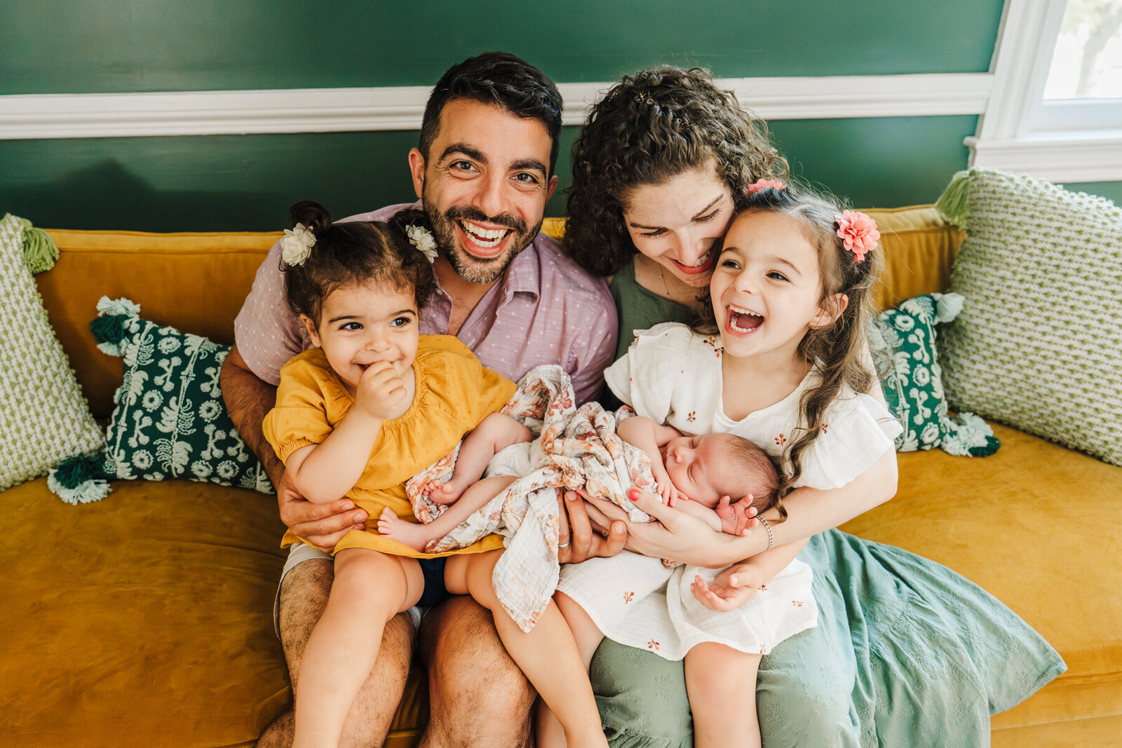 family laughs together on a mustard couch in a green room holding new baby
