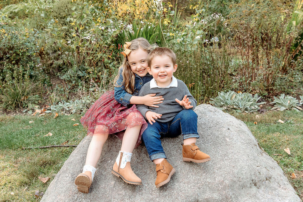 Sweet sibling portrait by Connecticut Family Photographer shows young brother and sister embracing while sitting on a large rock in a natural garden setting