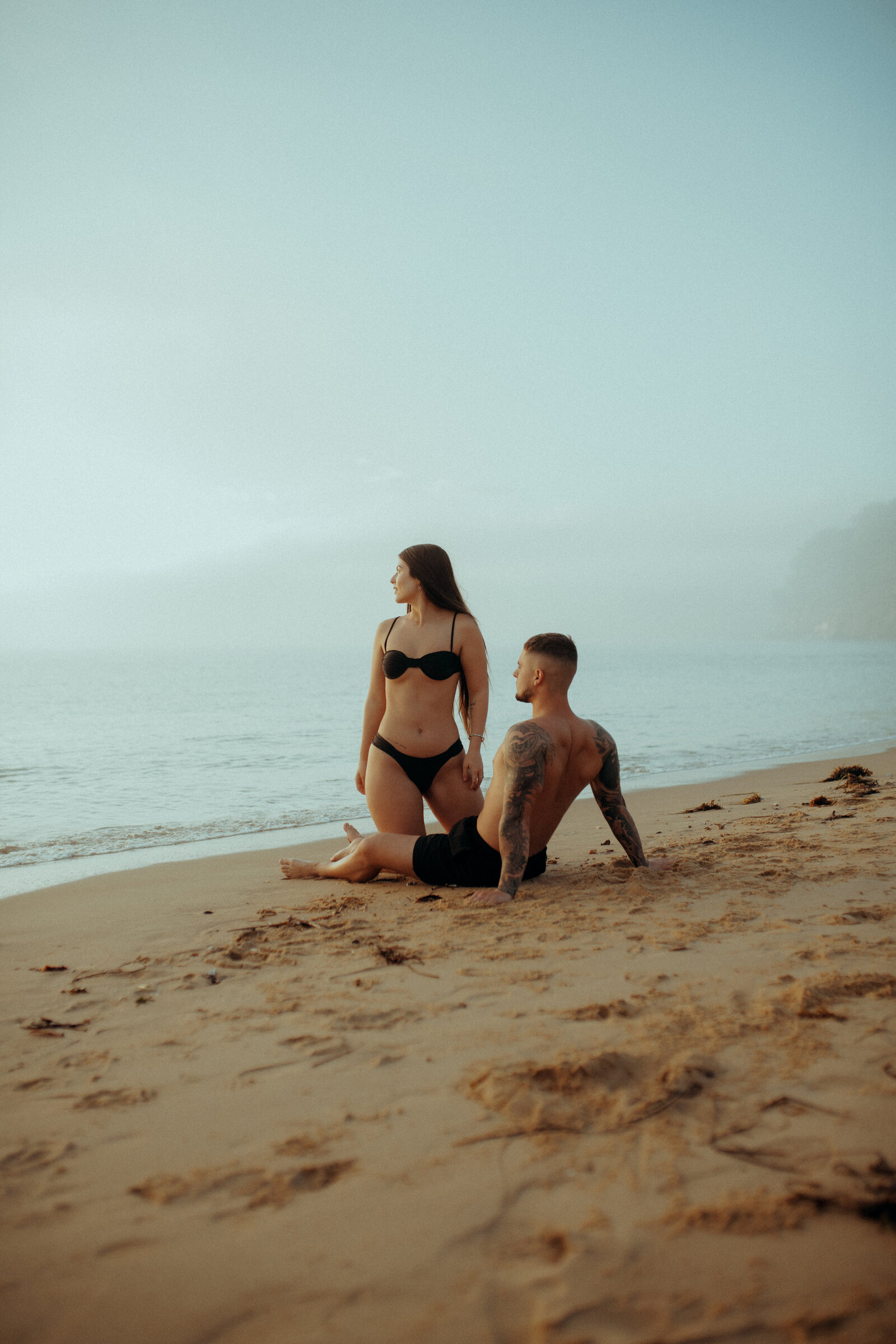 A woman in a black bikini stands on a sandy beach beside a seated, tattooed man facing the ocean on a foggy day.