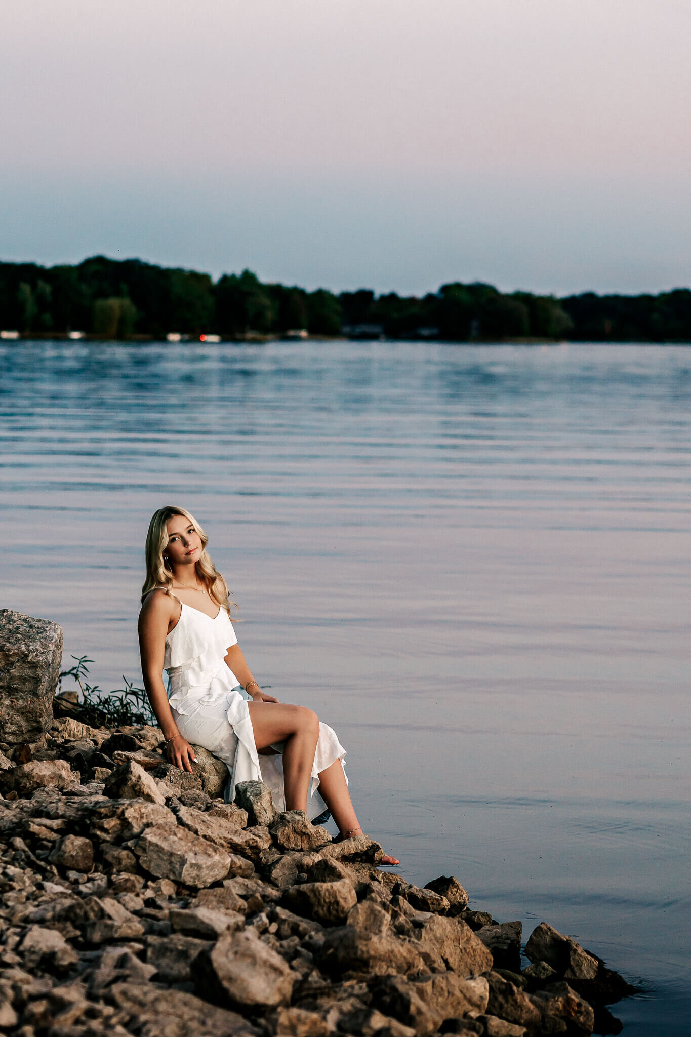 A woman in a white dress sits on rocky terrain beside a calm lake. The background shows a line of trees under a clear sky at dusk.