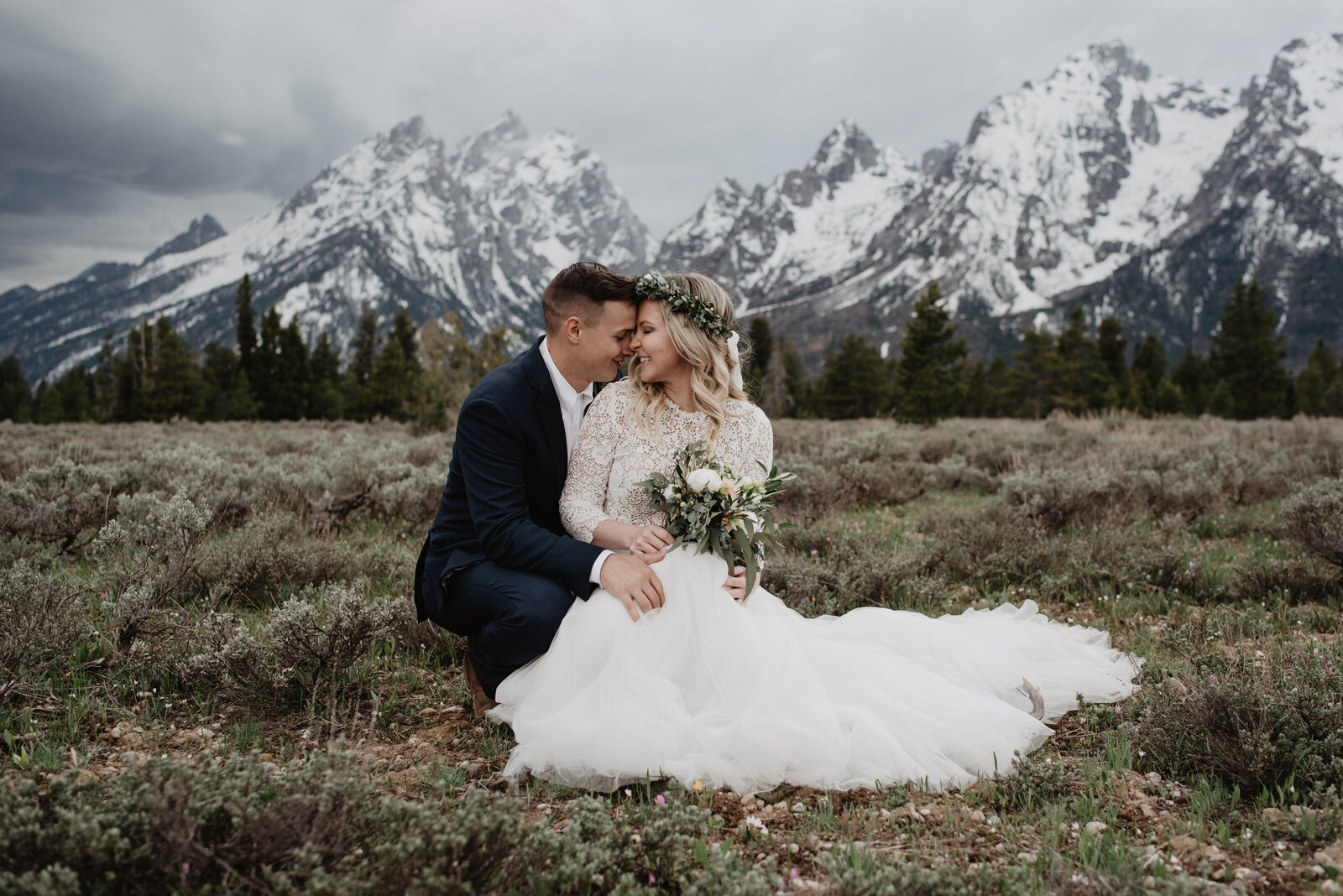 wedding photos in a meadow in front of the tetons with bride and groom sitting together and leaning in to rest their foreheads on one another with the mountains covered in snow in the distance with a storm rolling in past the mountains