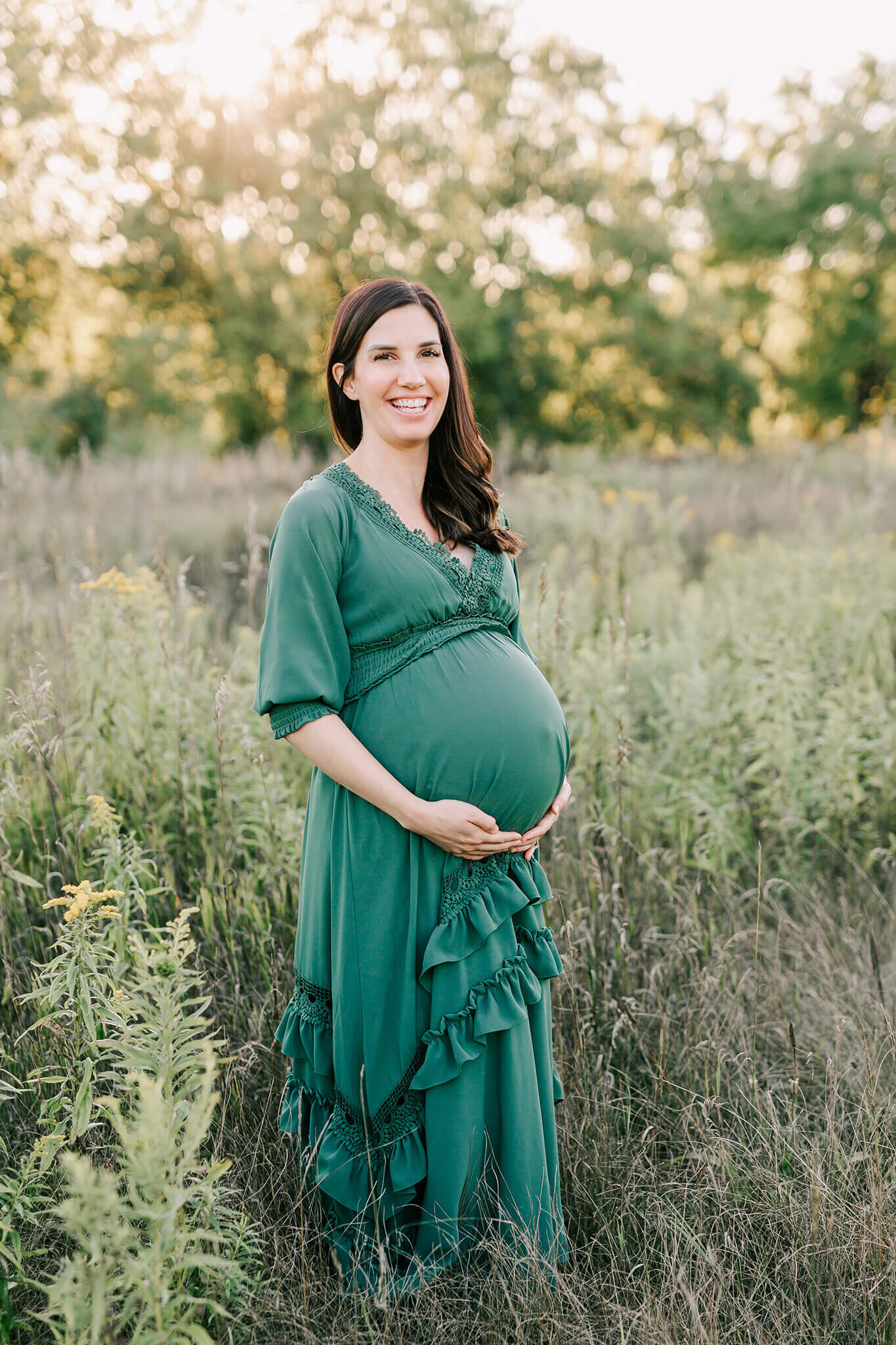 Expectant mother in a green dress stands in a field and smiles excitedly at the camera