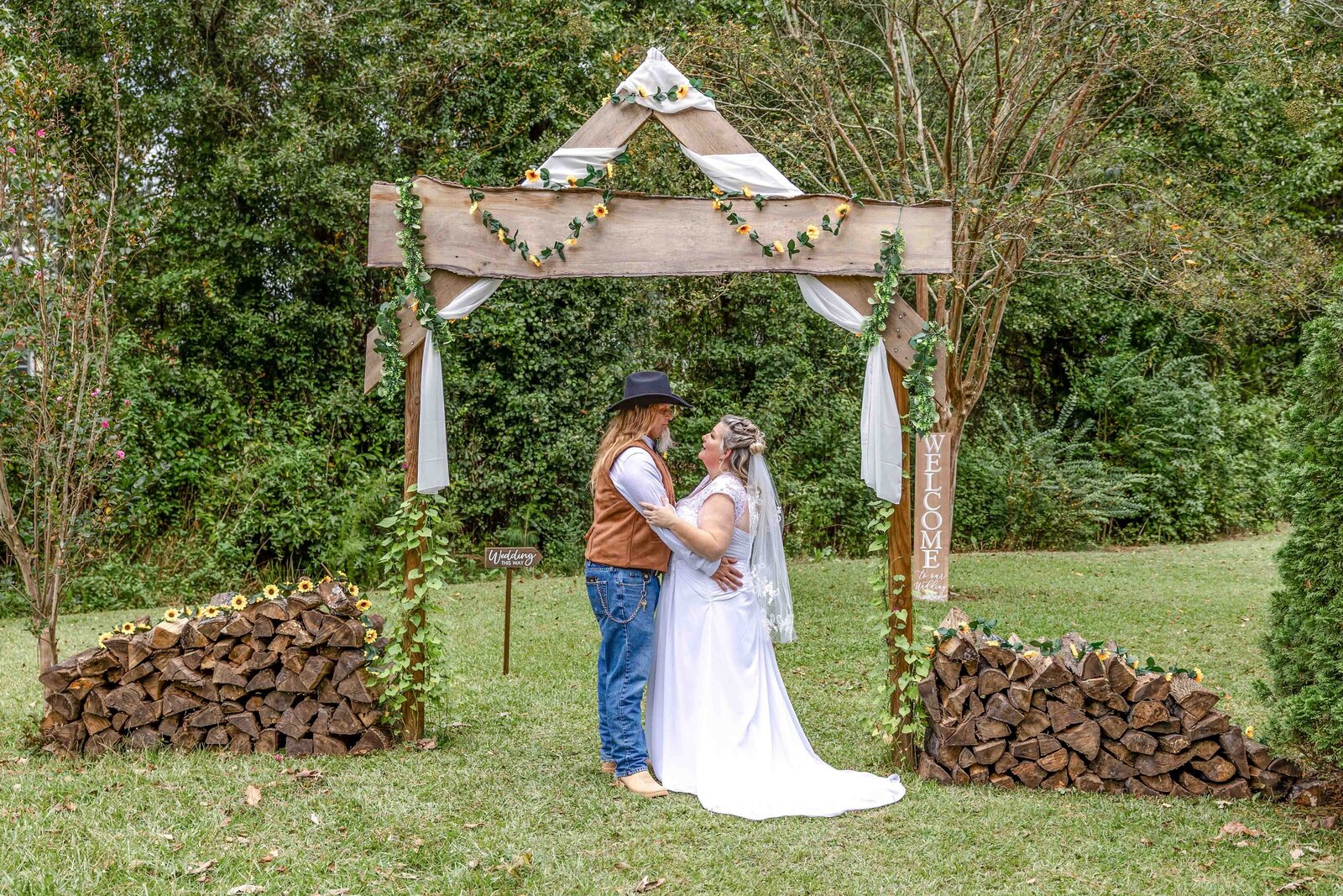 The bride and groom stand together under a rustic wooden ceremony arbor adorned with sunflowers and greenery, captured during a NC wedding, showcasing country charm and natural beauty.