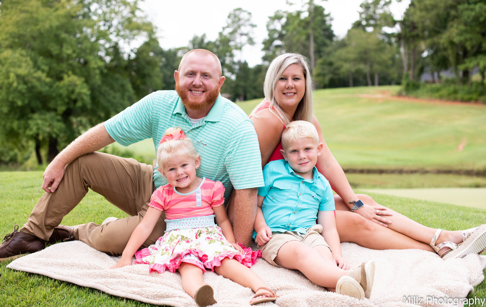 a mother, father, son, and daughter outdoors on a picnic blanket smiling at the camera photographed by Millz Photography in Greenville, SC