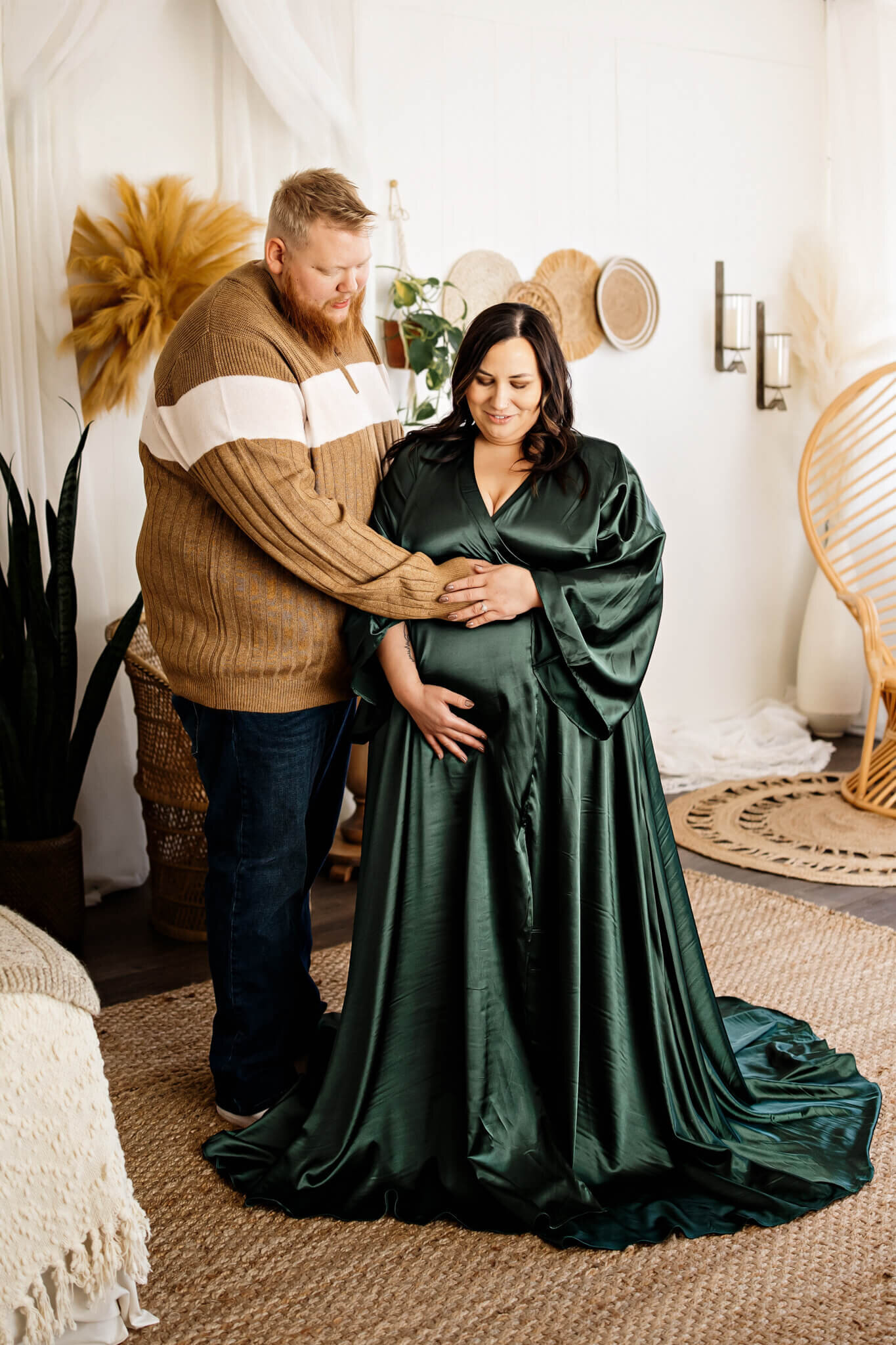 A smiling couple poses indoors for beautiful maternity portraits; the woman wears a flowing dark green gown and cradles her pregnant belly while the man gently rests his hand on her bump, captured by a Bloomington maternity photographer amid warm, plant-filled décor.
