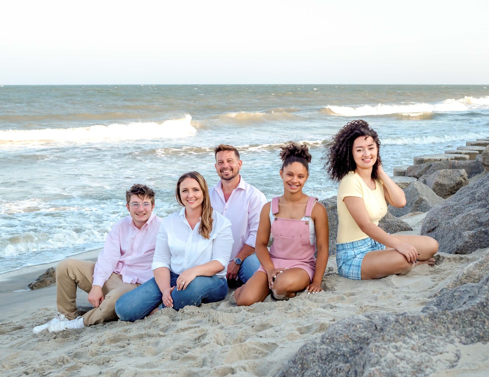 A group of friends sits casually on the rocky shoreline, dressed in pastel and casual attire. The ocean waves and breeze create a relaxed atmosphere. This image showcases affordable family photography in Wilmington, NC, capturing candid and joyful moments.