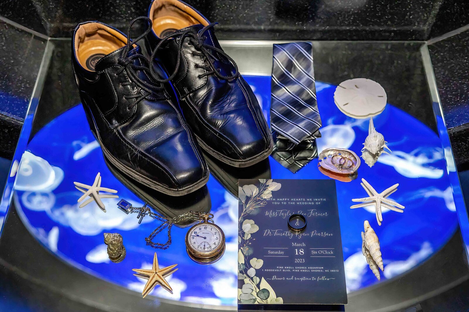 A flat lay featuring polished black shoes, a striped tie, wedding invitations, starfish, and nautical accents, captured on a glowing surface during a Pine Knoll Shores, NC wedding, blending elegance and coastal charm.
