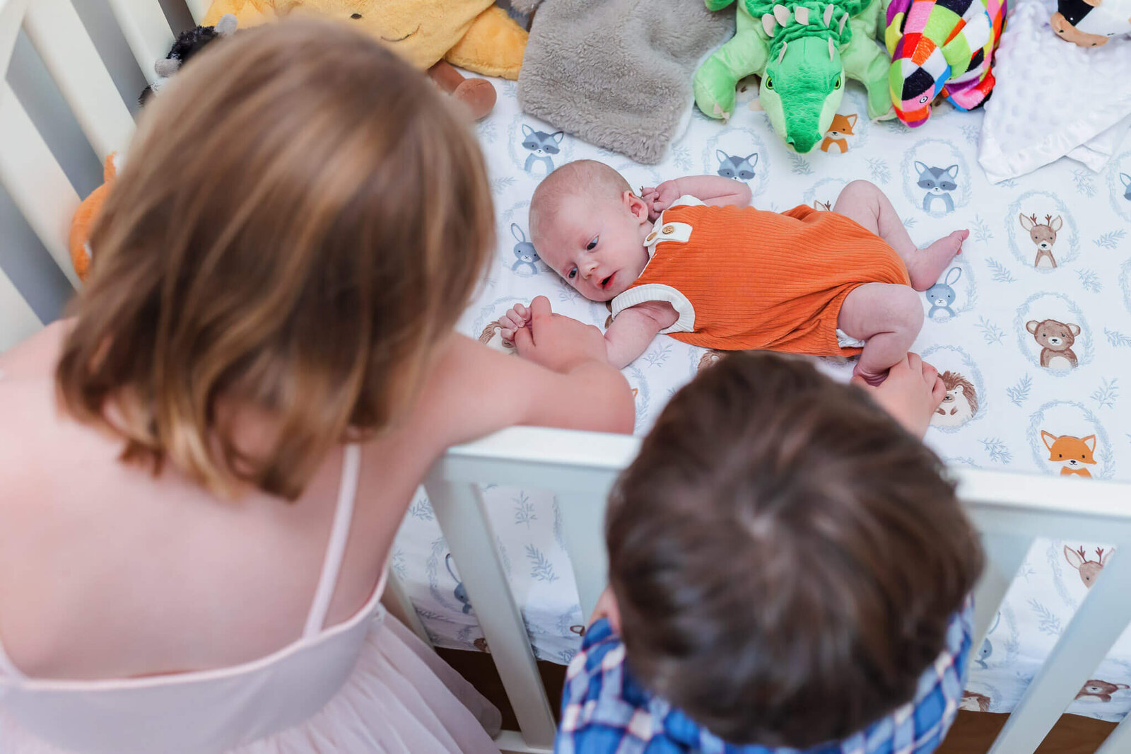 A newborn boy laying in his crib while his siblings look into the crib at him.