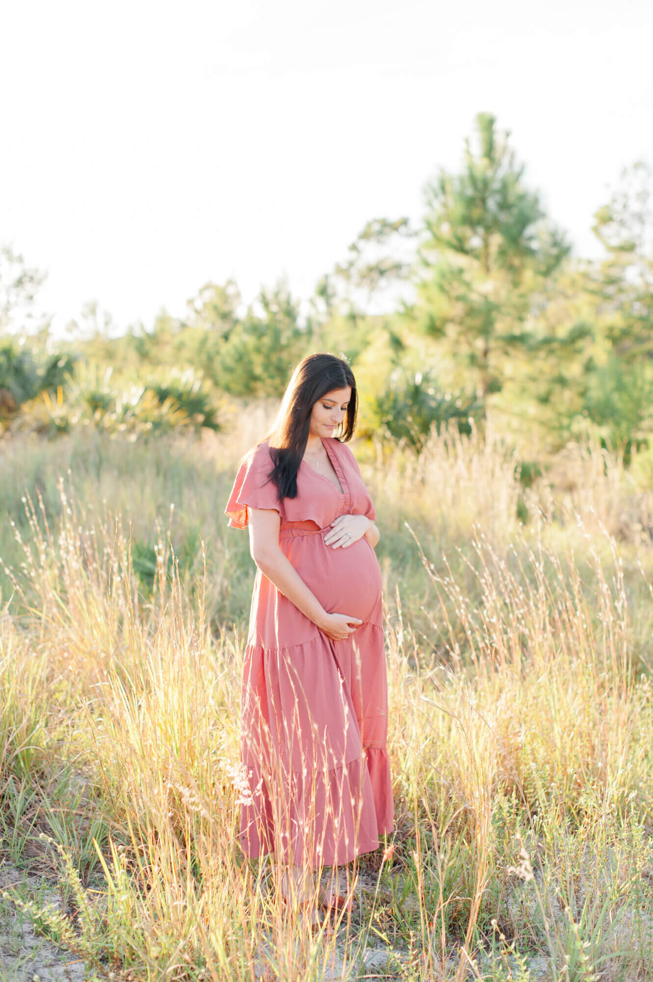 Beautiful new mother stands in a tall grass field wearing a pink dress during golden hour