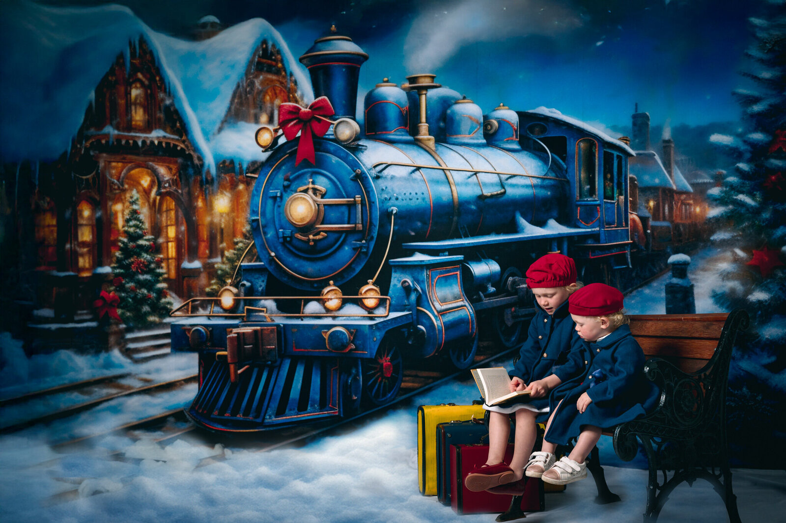 Maeve and Marcie in blue coats and red hats reading a book infront of a Christmas train scene in the snow, sitting on a park bench