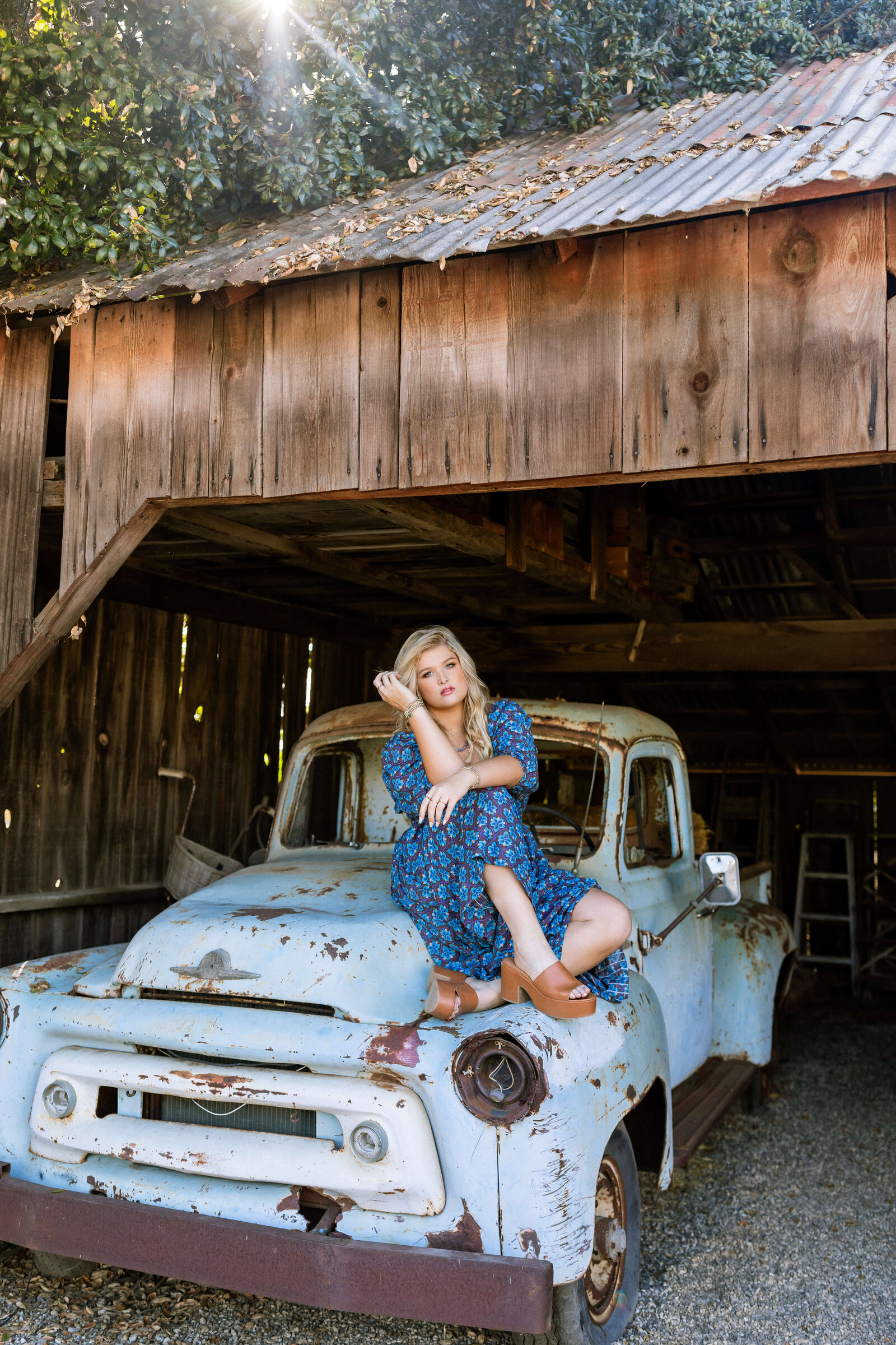 blonde high school girl wearing a blue dress with brown heels posing on a blue rusted car under a barn