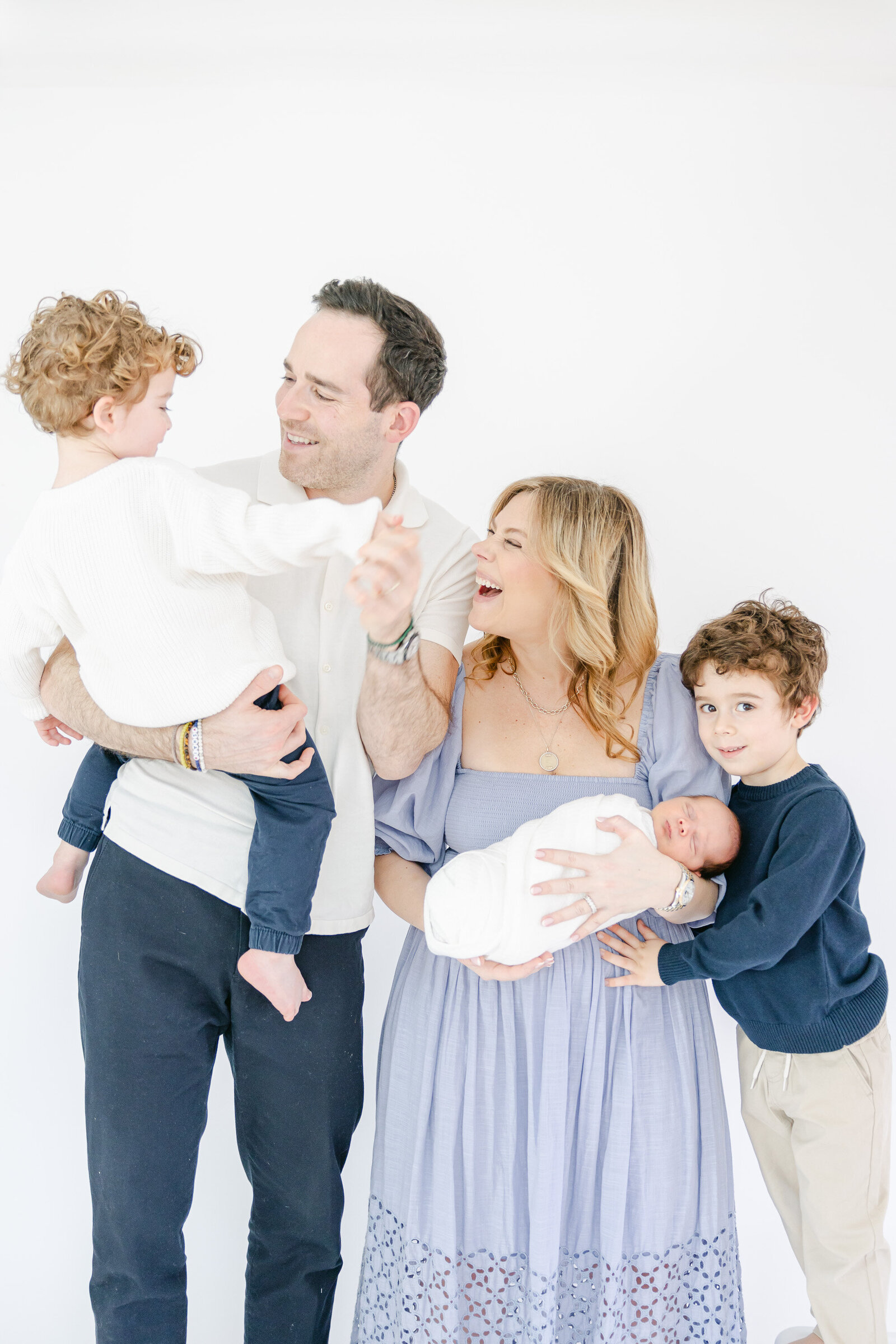 A cheerful family of five shares a candid moment during an in-home lifestyle photography session in Fairfield County, Connecticut. The mother in a soft periwinkle dress cradles a newborn while two young boys and their father laugh and interact naturally, captured in a bright white studio setting by Nicole Neira Photography.