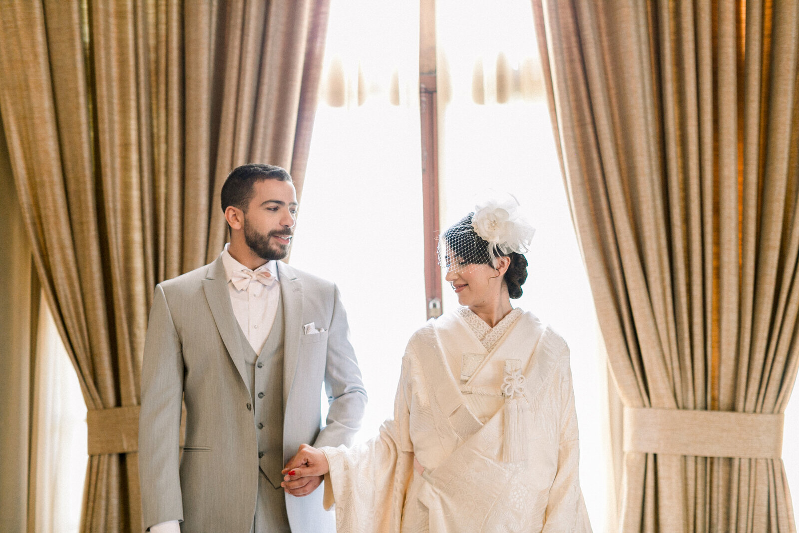 Bride and groom holding hands in white shiromuku by tall curtains at The Ebell Club Long Beach, Los Angeles California. Perfect for wedding kimono rental in Los Angeles | ロサンゼルスのエベルクラブでカーテン前に手をつなぐ白無垢姿の新郎新婦。着物レンタル可能。