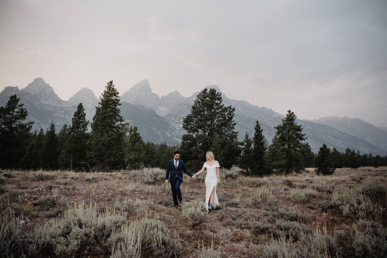 Grand Teton wedding with bride and groom holding hands at dusk as they walk together in a meadow with a forest behind them for a moody wedding day