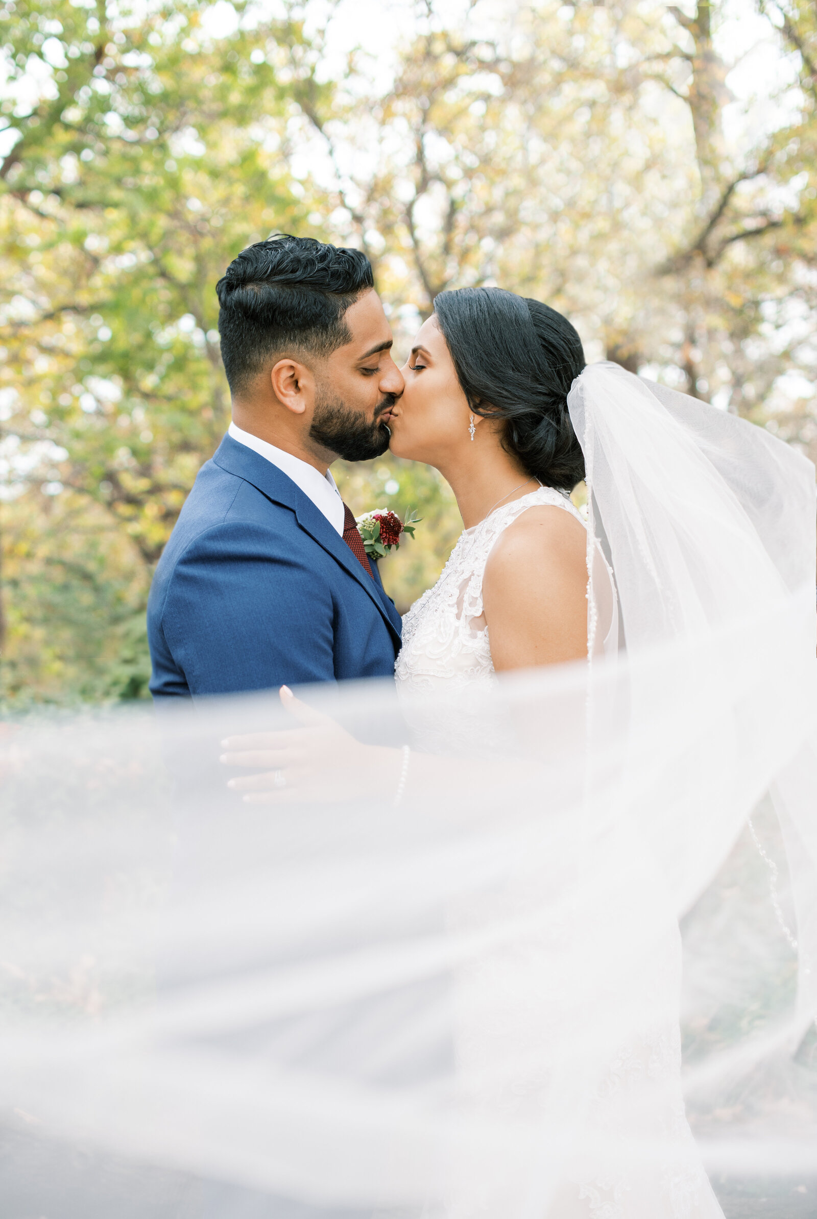 Portrait of a bride and groom in a white wedding gown and blue tuxedo kissing with large trees outback.