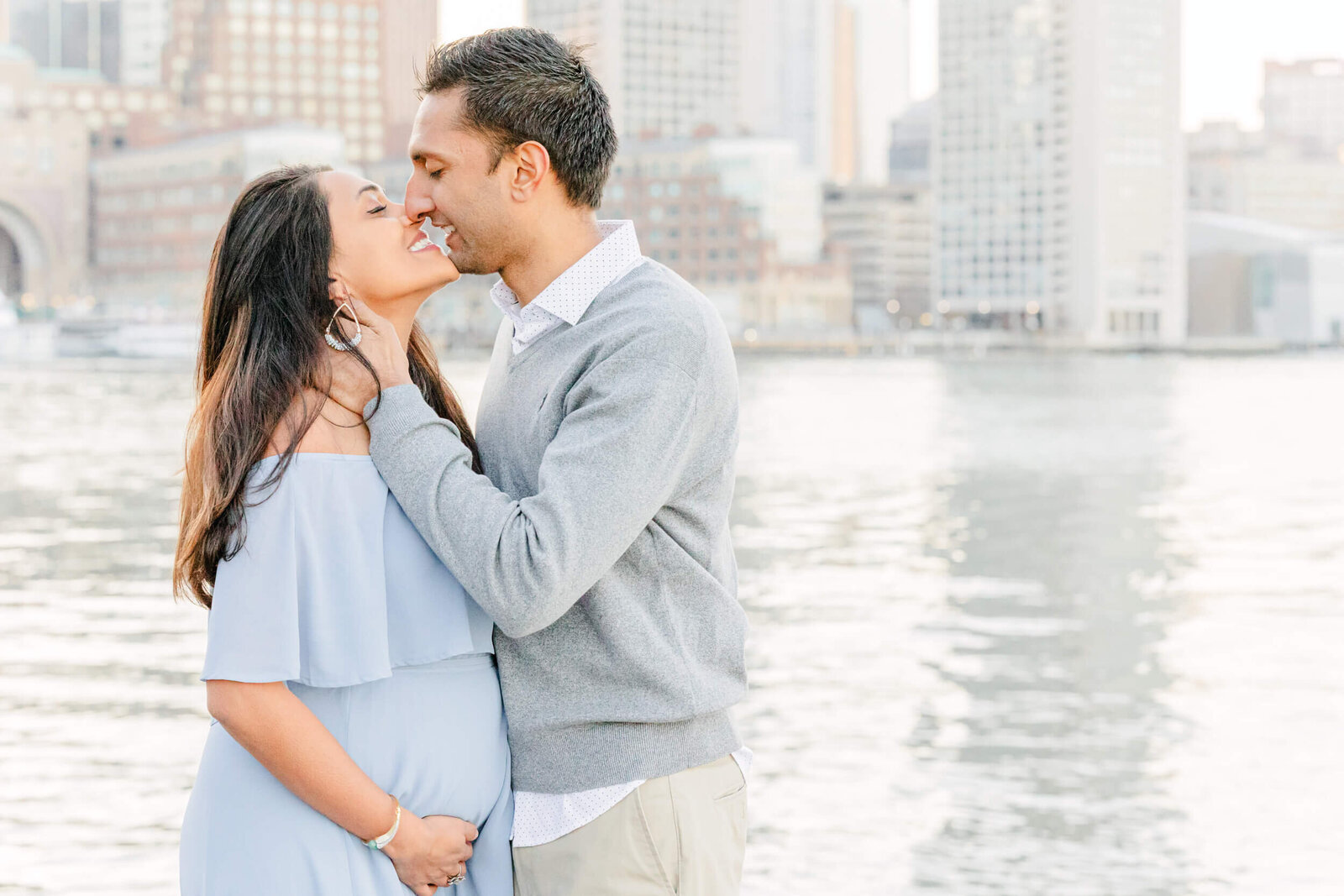 Pregnant woman and her husband lean in for a kiss in front of Boston harbor with the city in the background