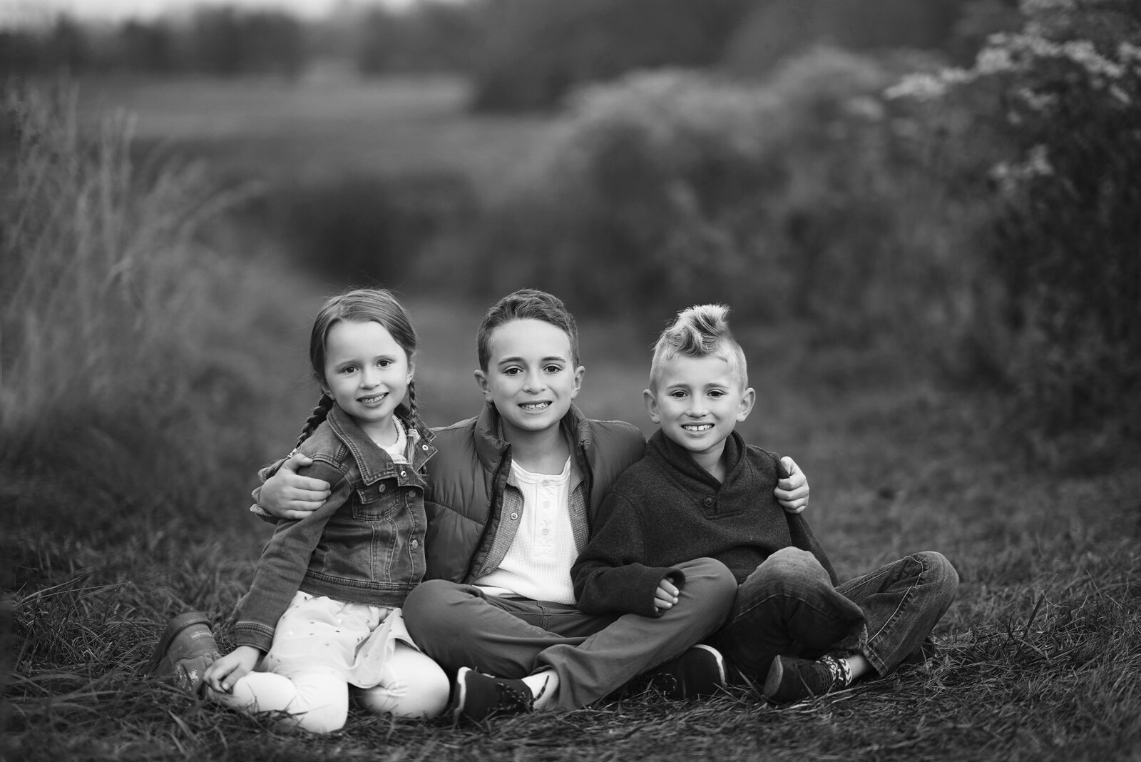 Big bother is sitting in a filed with his arms around his little brother and little sister who are sitting beside him. All are smiling at the camera. Image captured by top Philadelphia and Main Line family photographer.
