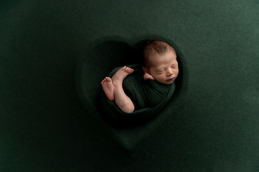 Newborn baby sleeps in a heart bowl on a green background.
